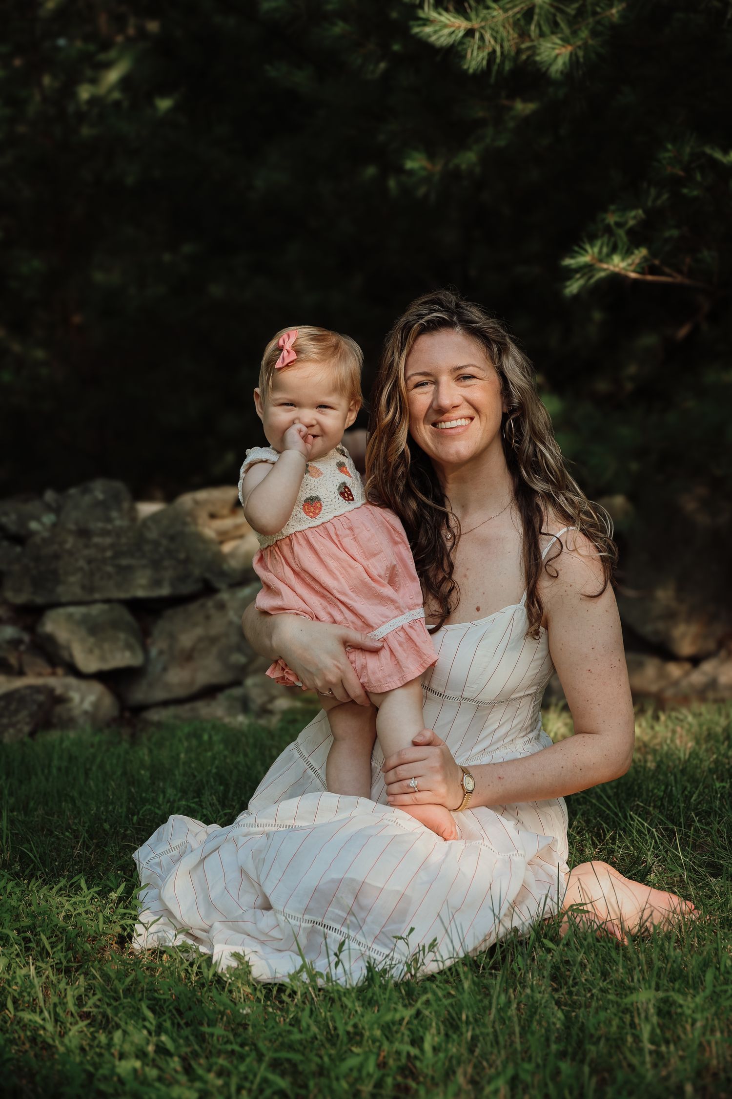 A tender moment between mother and child sitting together on grass in white dresses during an outdoor photo session.