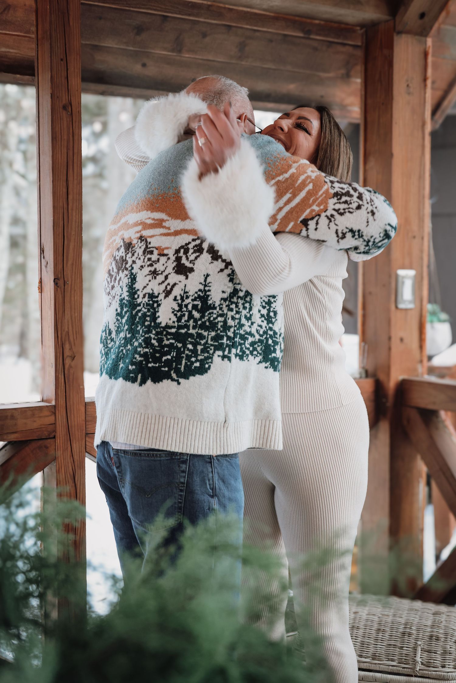 Two people wearing matching white winter sweaters with green pine tree patterns embrace in a rustic wooden porch setting.