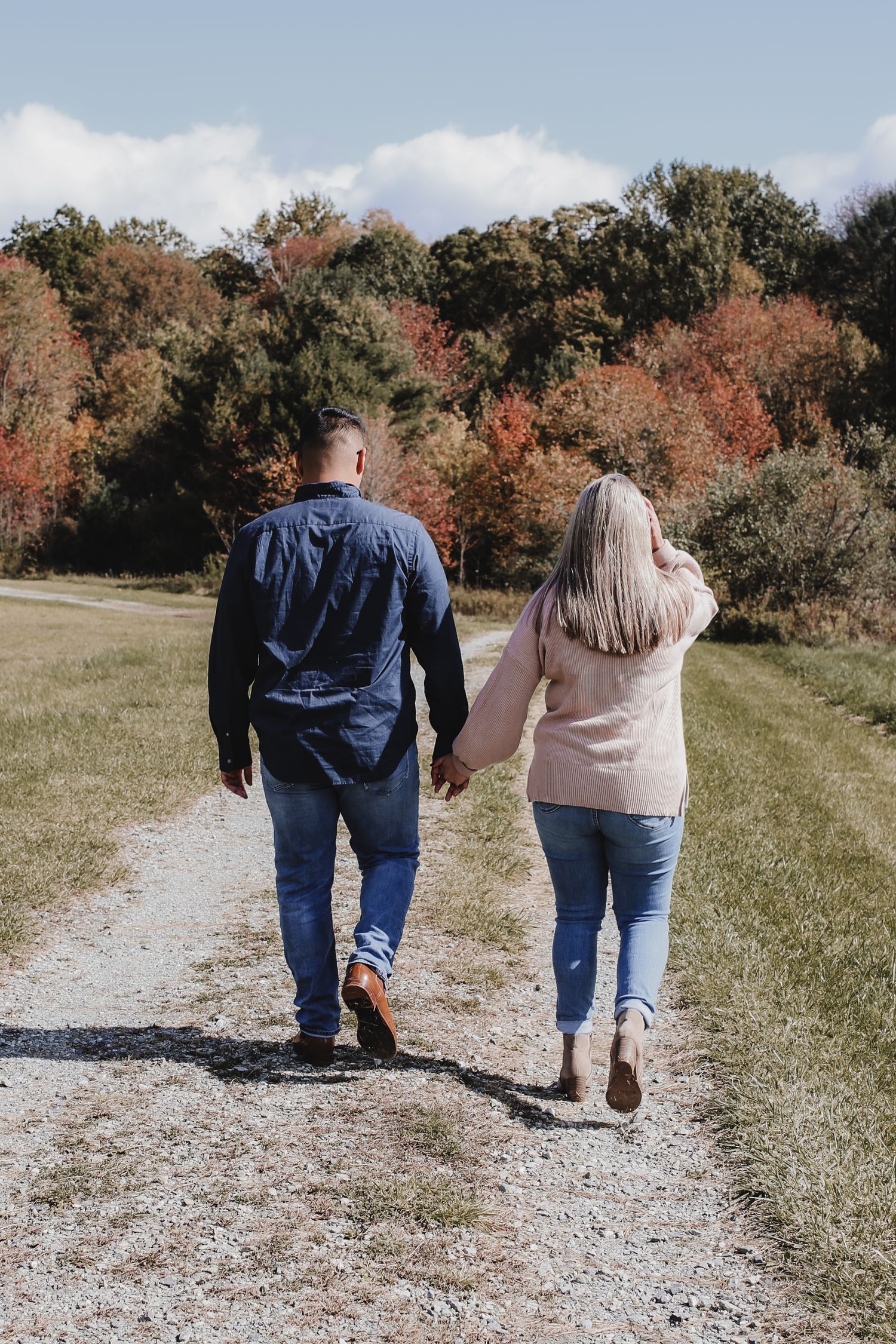 A couple walking hand in hand on a gravel path surrounded by colorful autumn trees and foliage.