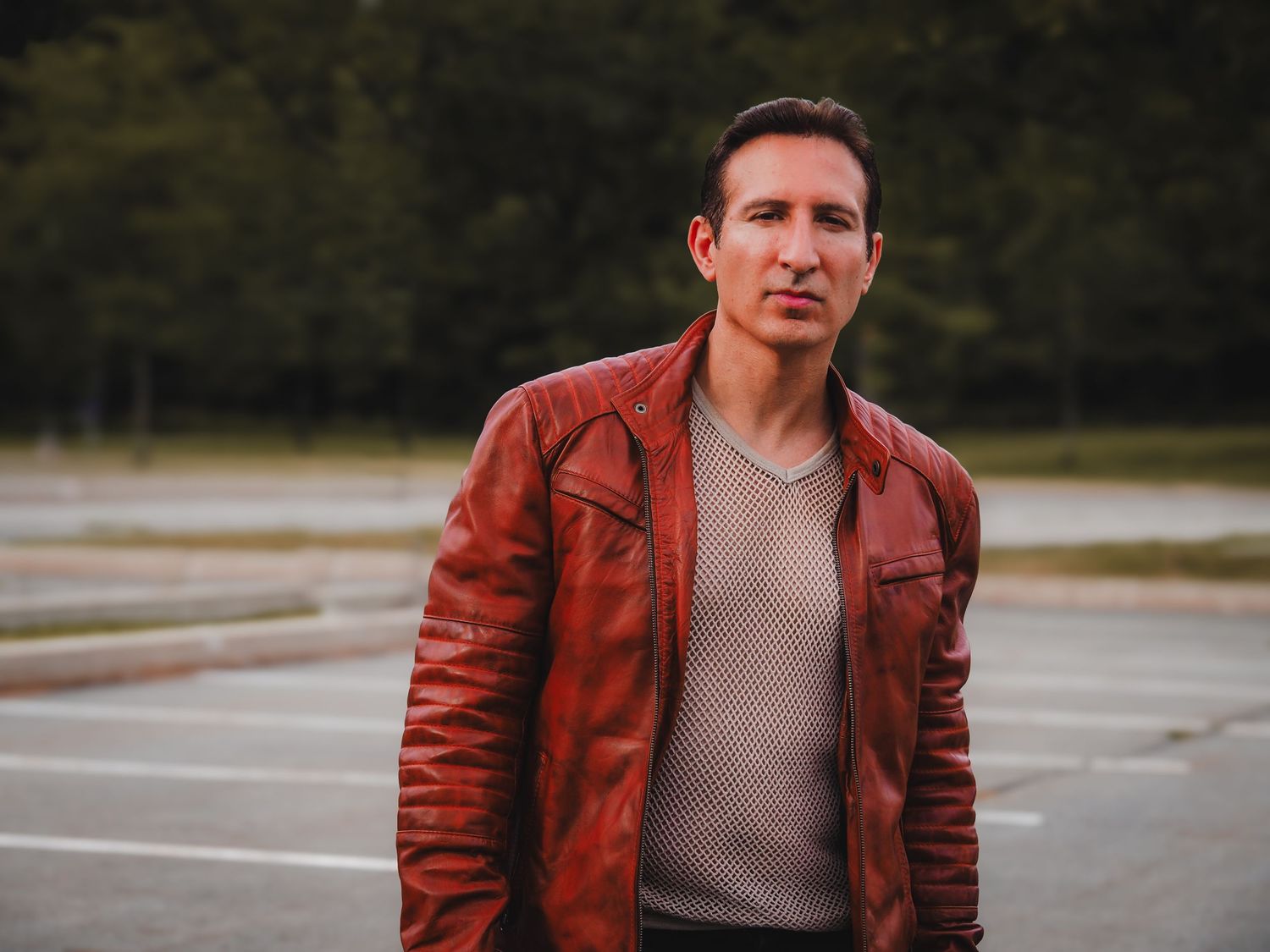 Person wearing a brown leather jacket and gray sweater stands in an empty parking lot on an overcast day.