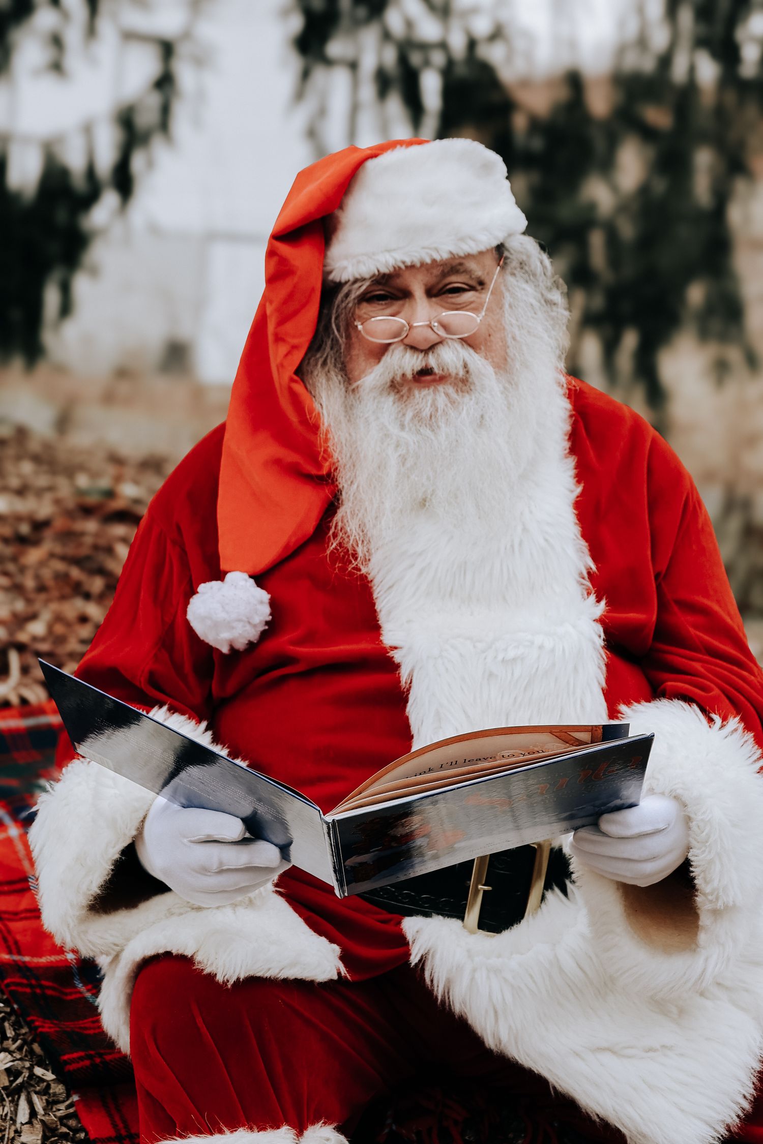 A figure in a traditional red and white holiday costume sits outdoors reading a book among autumn leaves.
