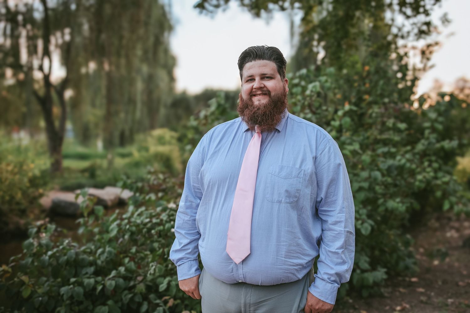 A person in a light blue dress shirt and pink tie stands in a lush outdoor garden setting.