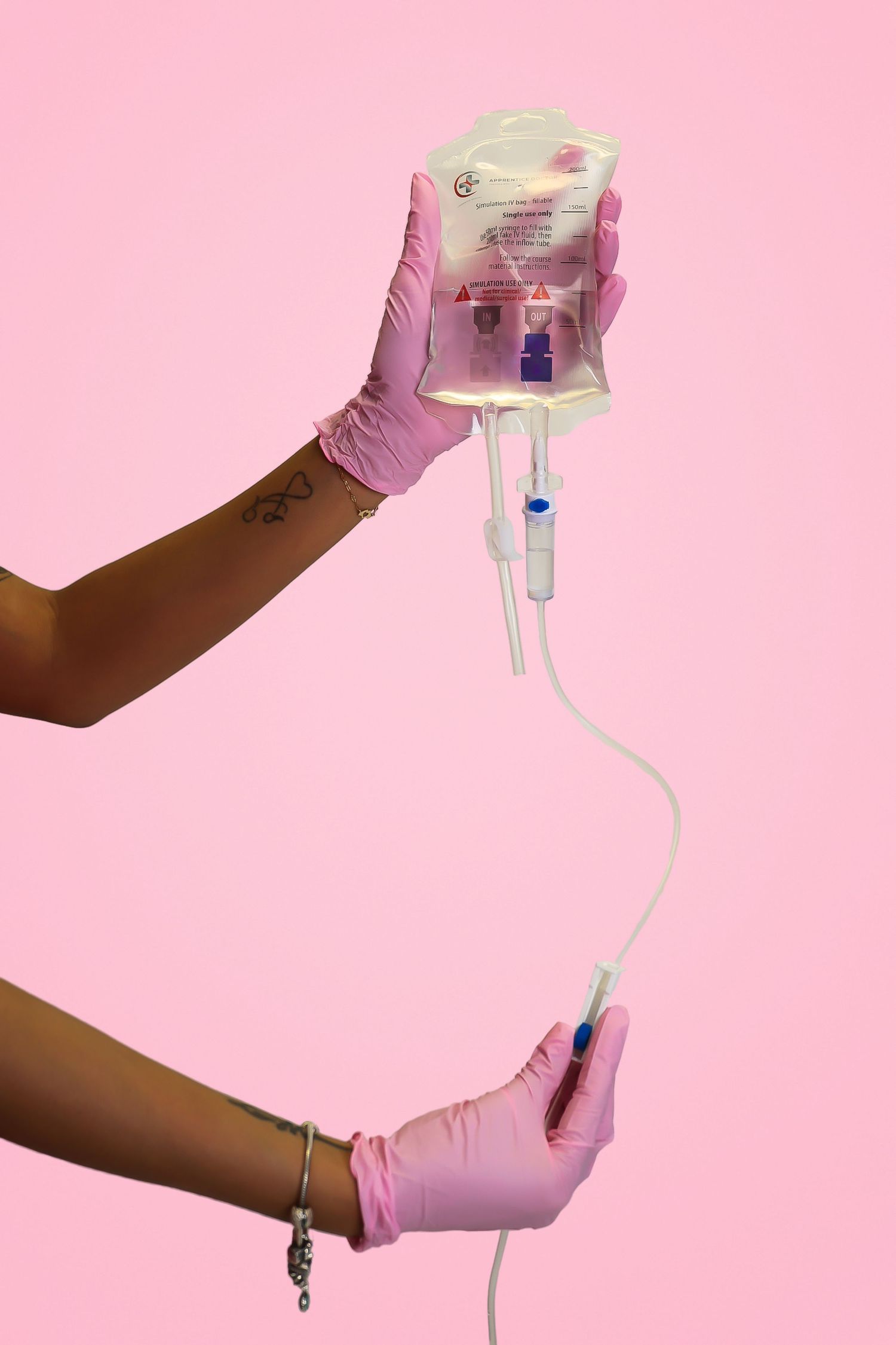 Hands in pink latex gloves holding an IV drip bag with tubing against a pink background.