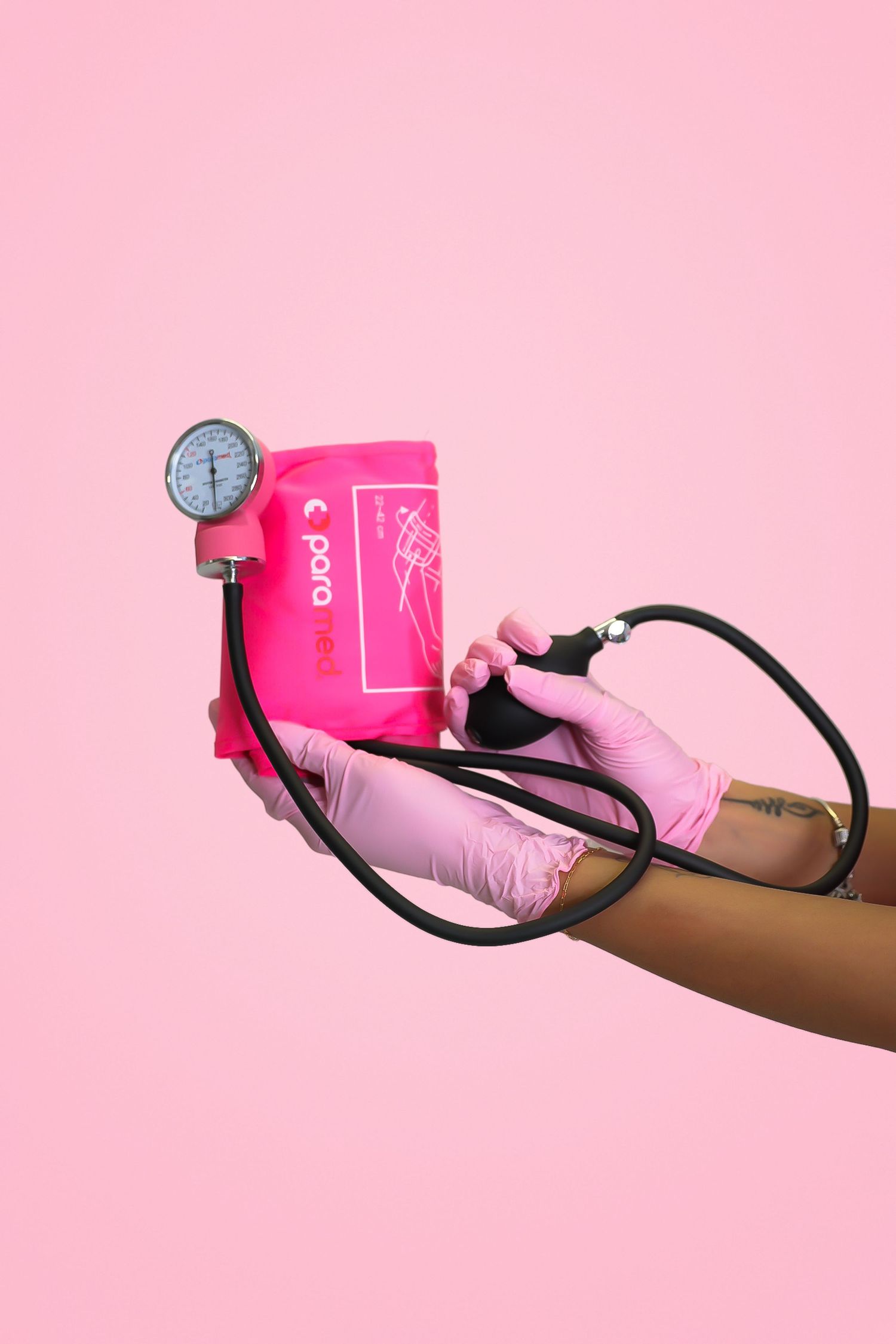 Hand in pink glove holding a bright pink blood pressure cuff and bulb on a pink background.