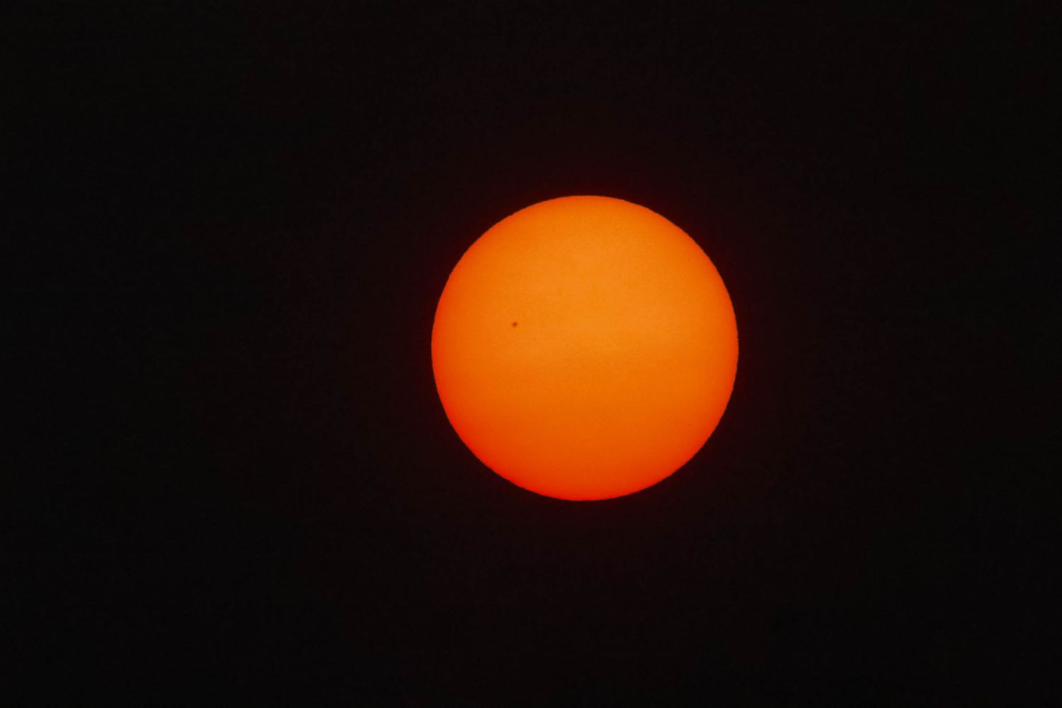 Bright orange sun with visible sunspots against a dark black sky, captured through a solar filter telescope.
