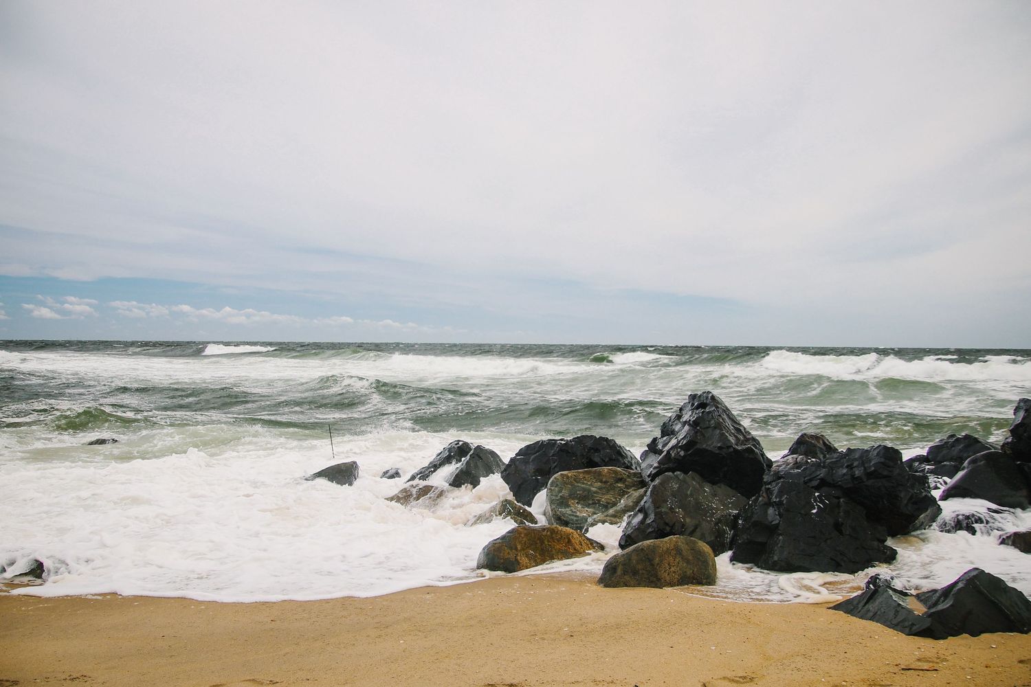 Rocky shoreline with crashing ocean waves on a sandy beach under a cloudy sky.