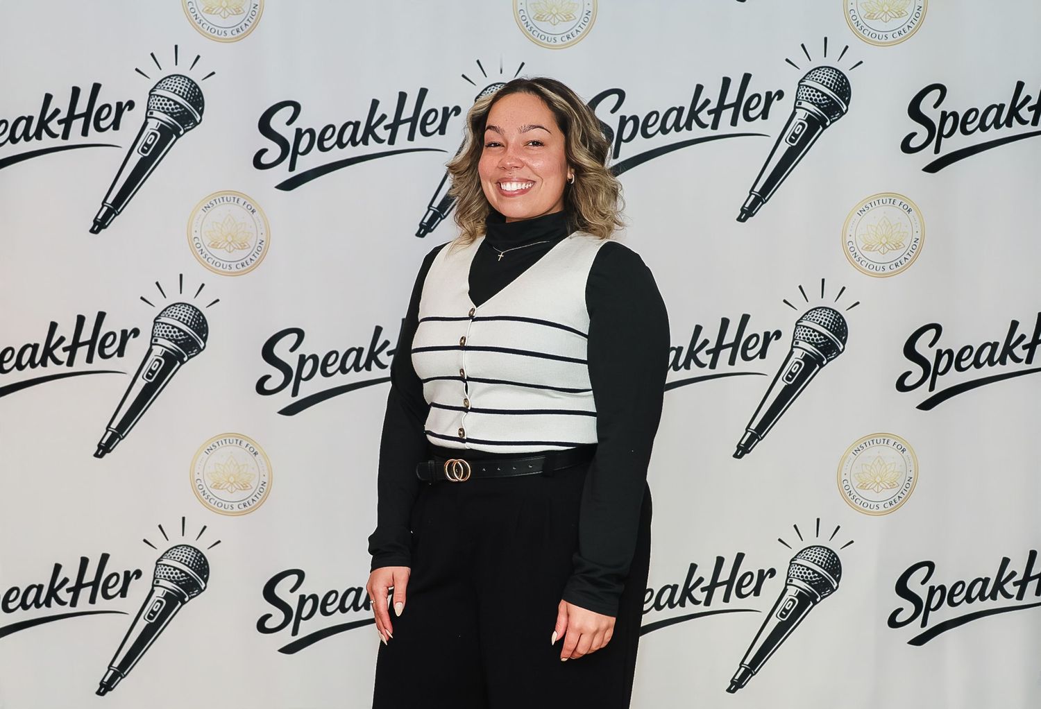Woman smiling in black and white outfit at SpeakHer event backdrop with microphone logos.