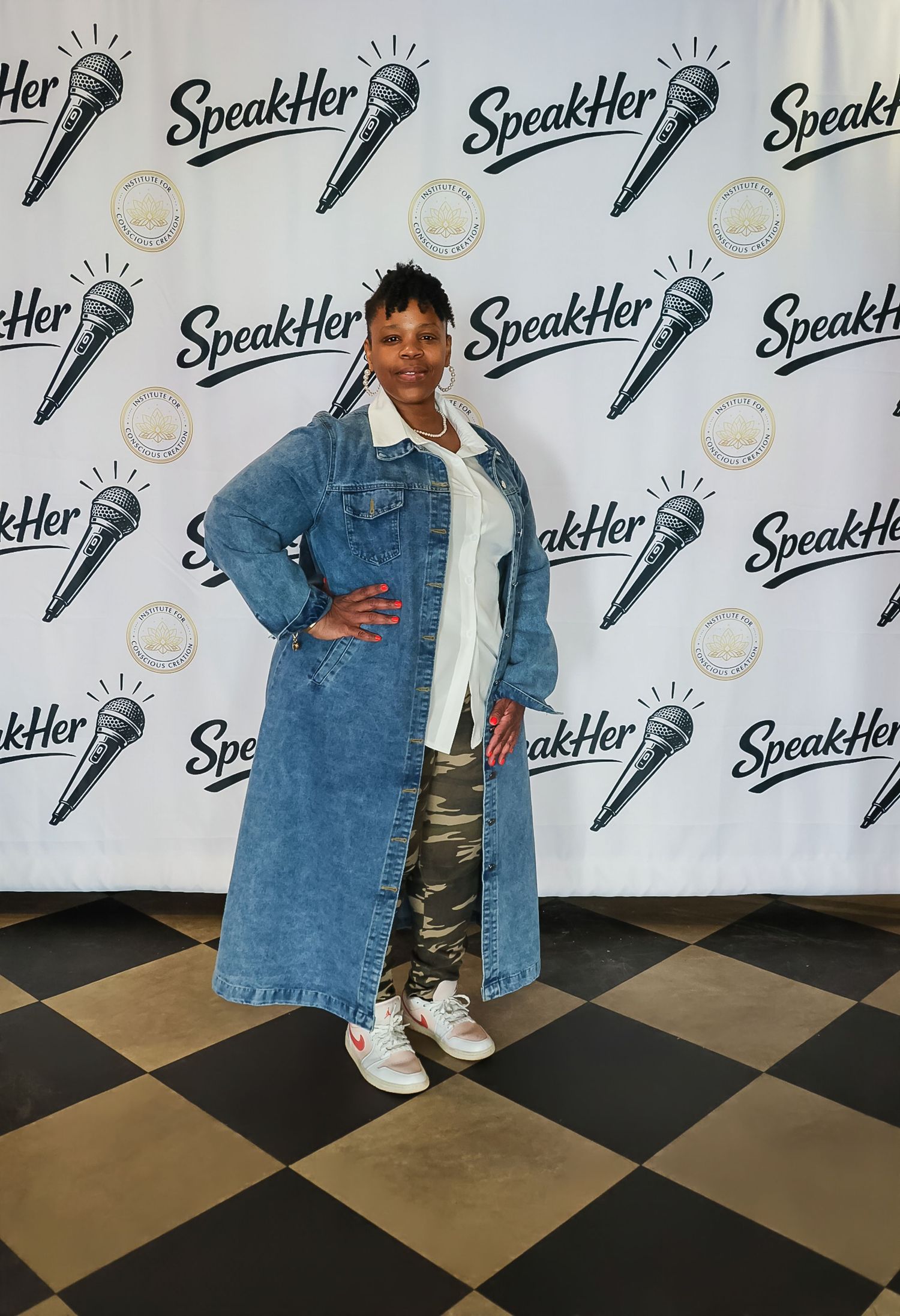 Woman in long denim coat and camo pants poses at SpeakHer event step-and-repeat backdrop.