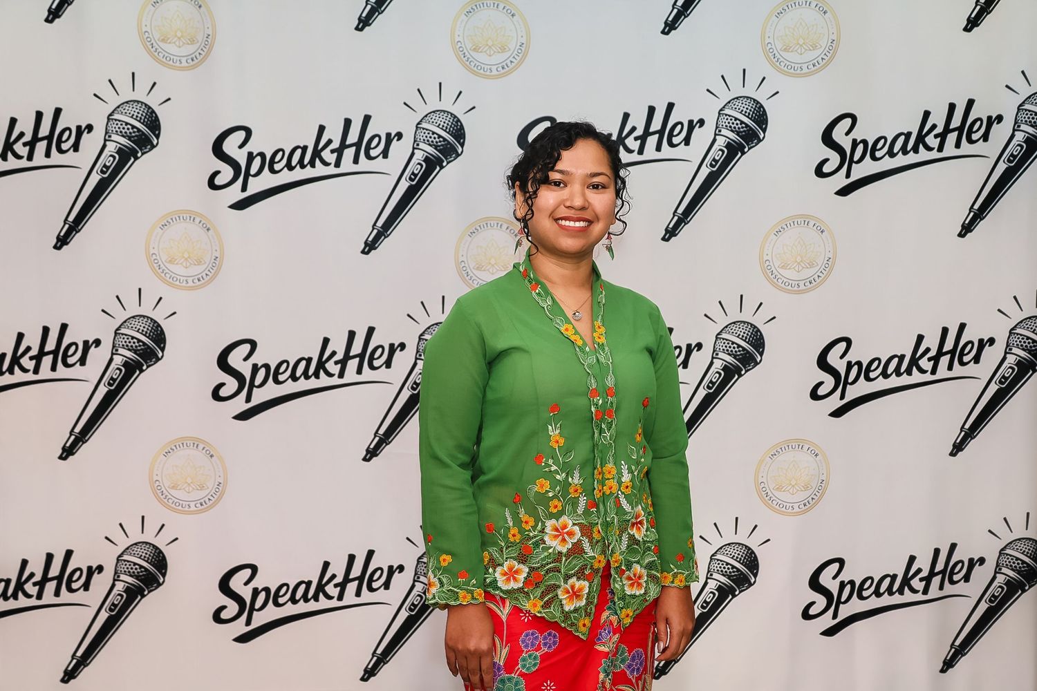 Woman in green floral top and pink skirt posing at SpeakHer event backdrop with microphone logos.