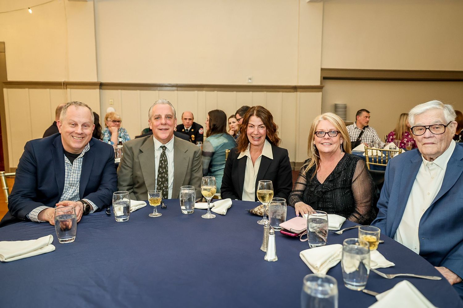 Five smiling guests seated at a banquet table with blue tablecloth, glasses, and formal dinnerware at an indoor event.
