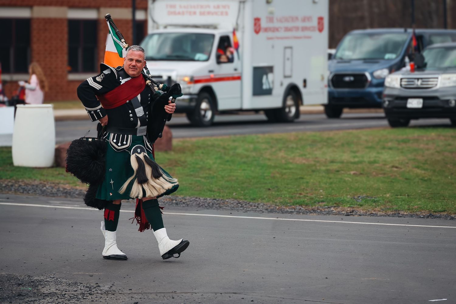 Scottish bagpiper in traditional kilt and regalia marching in an outdoor parade near parked vehicles.