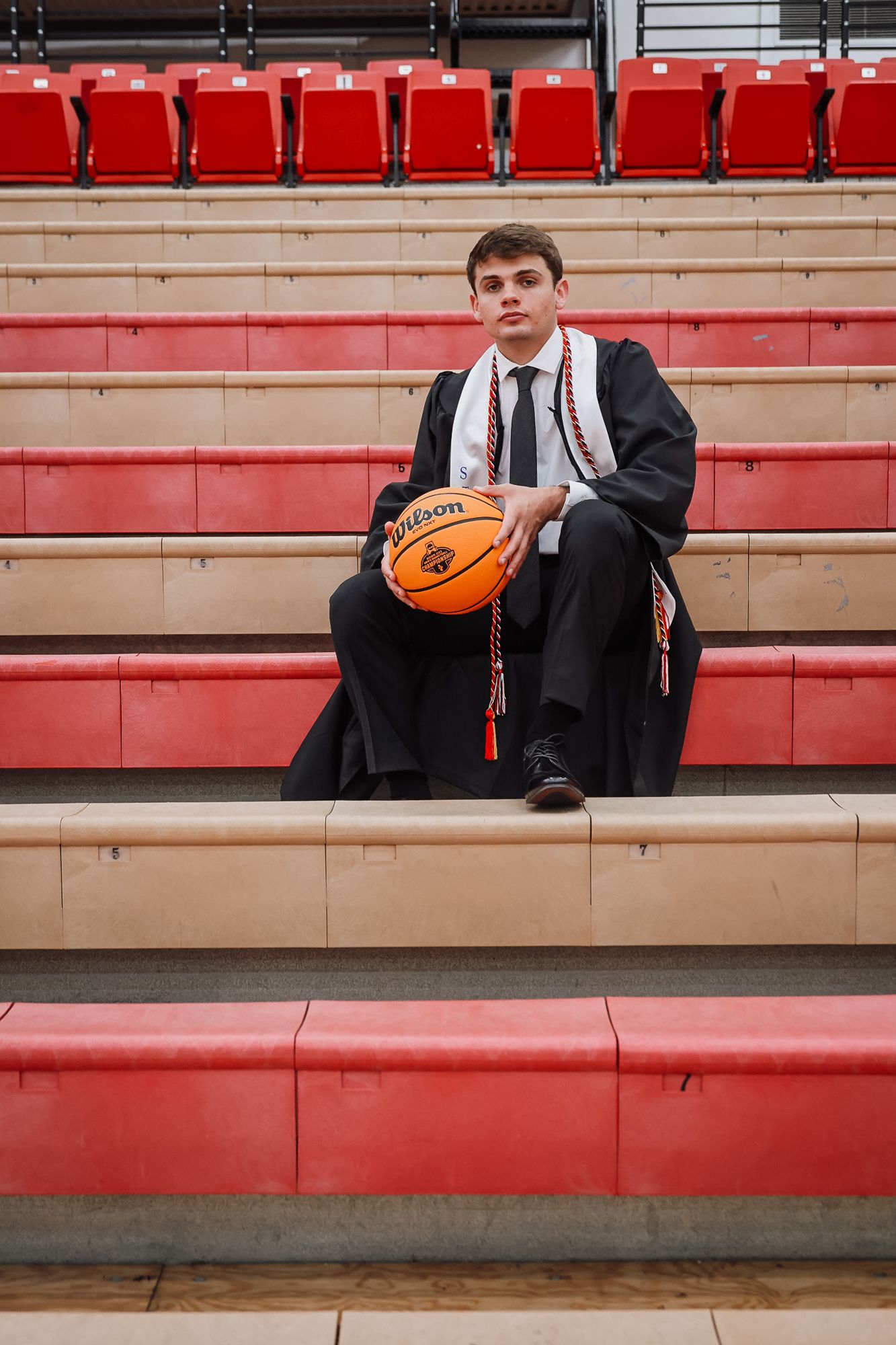 Young man in graduation gown holding a Wilson basketball while sitting in red bleachers at a gymnasium.
