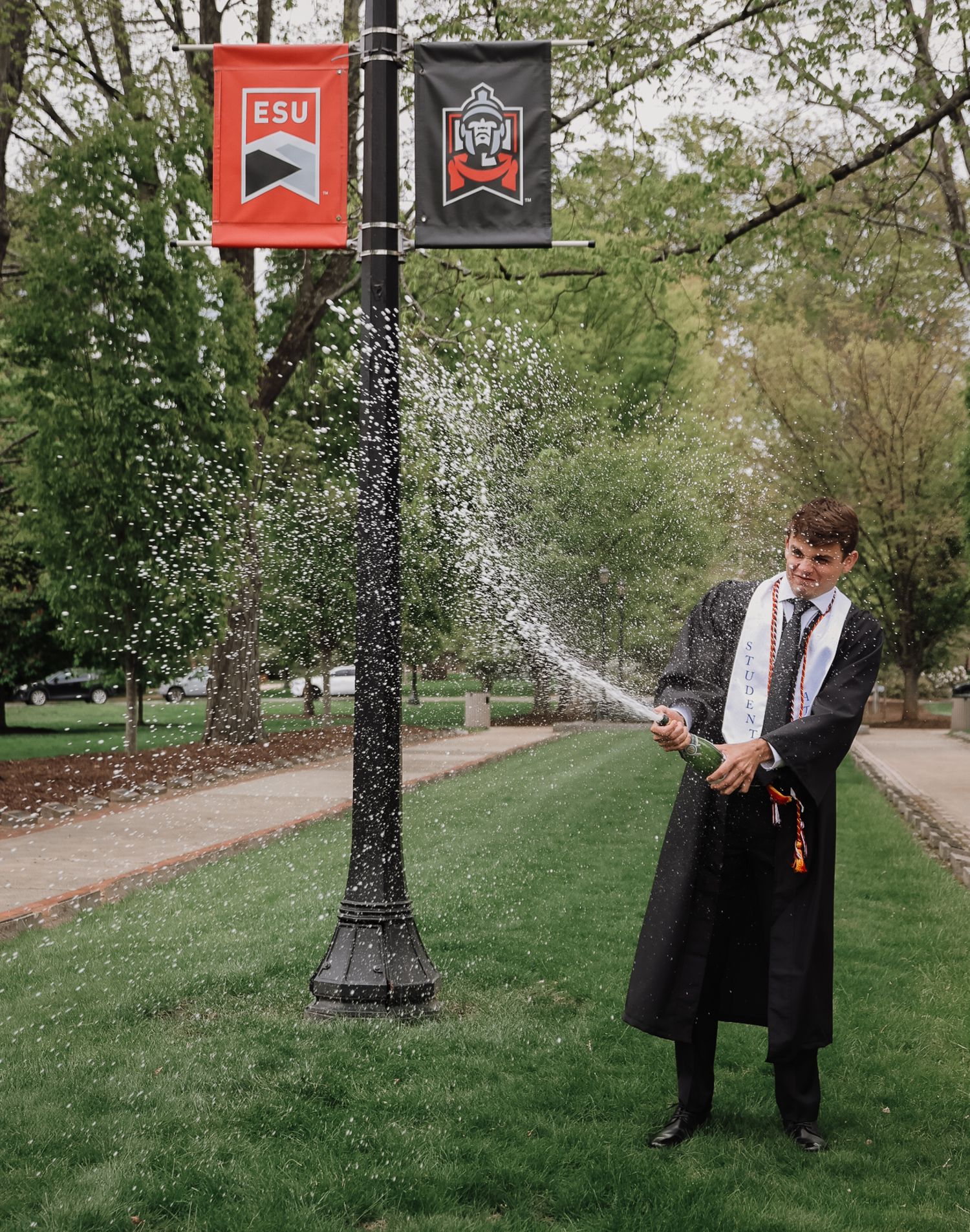 Graduate in cap and gown pops champagne bottle on ESU campus pathway surrounded by green trees.