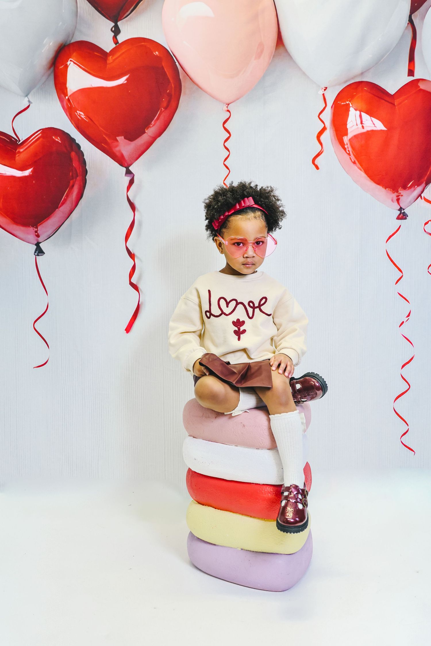 A young girl in a cream ‘love’ sweater sits on colorful stacked cushions surrounded by red and pink heart-shaped balloons.