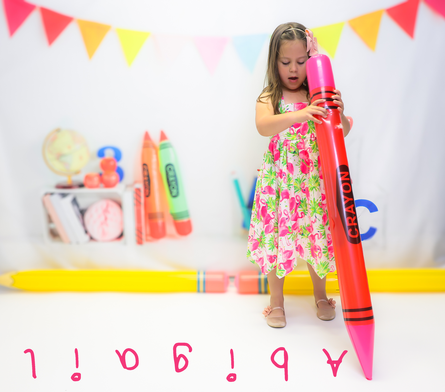 Young girl in floral dress holding oversized pink Crayola crayon with colorful school supplies backdrop and name Abigail below.