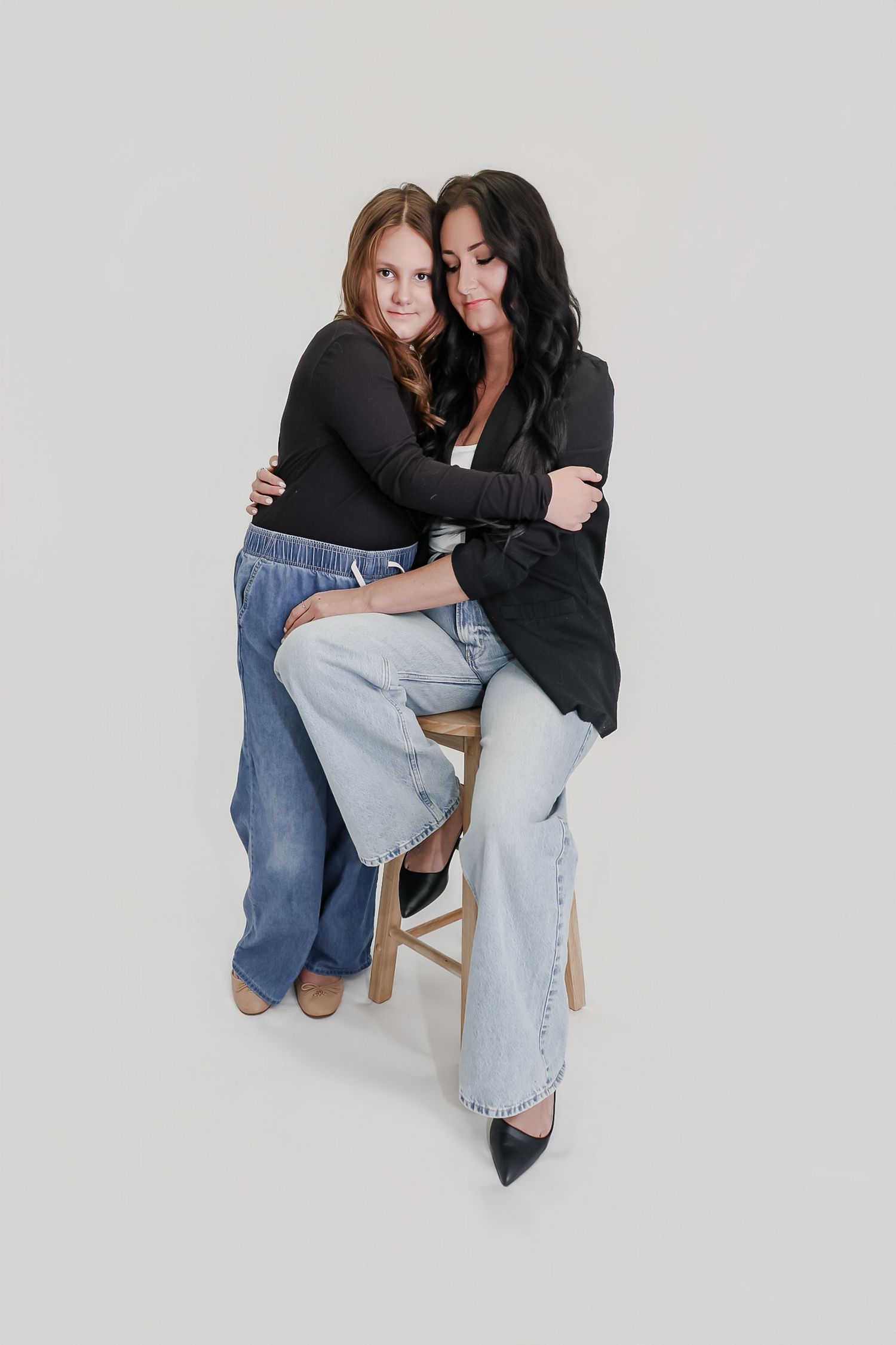 A woman and young girl in jeans and black tops share a warm embrace while sitting on a wooden stool.