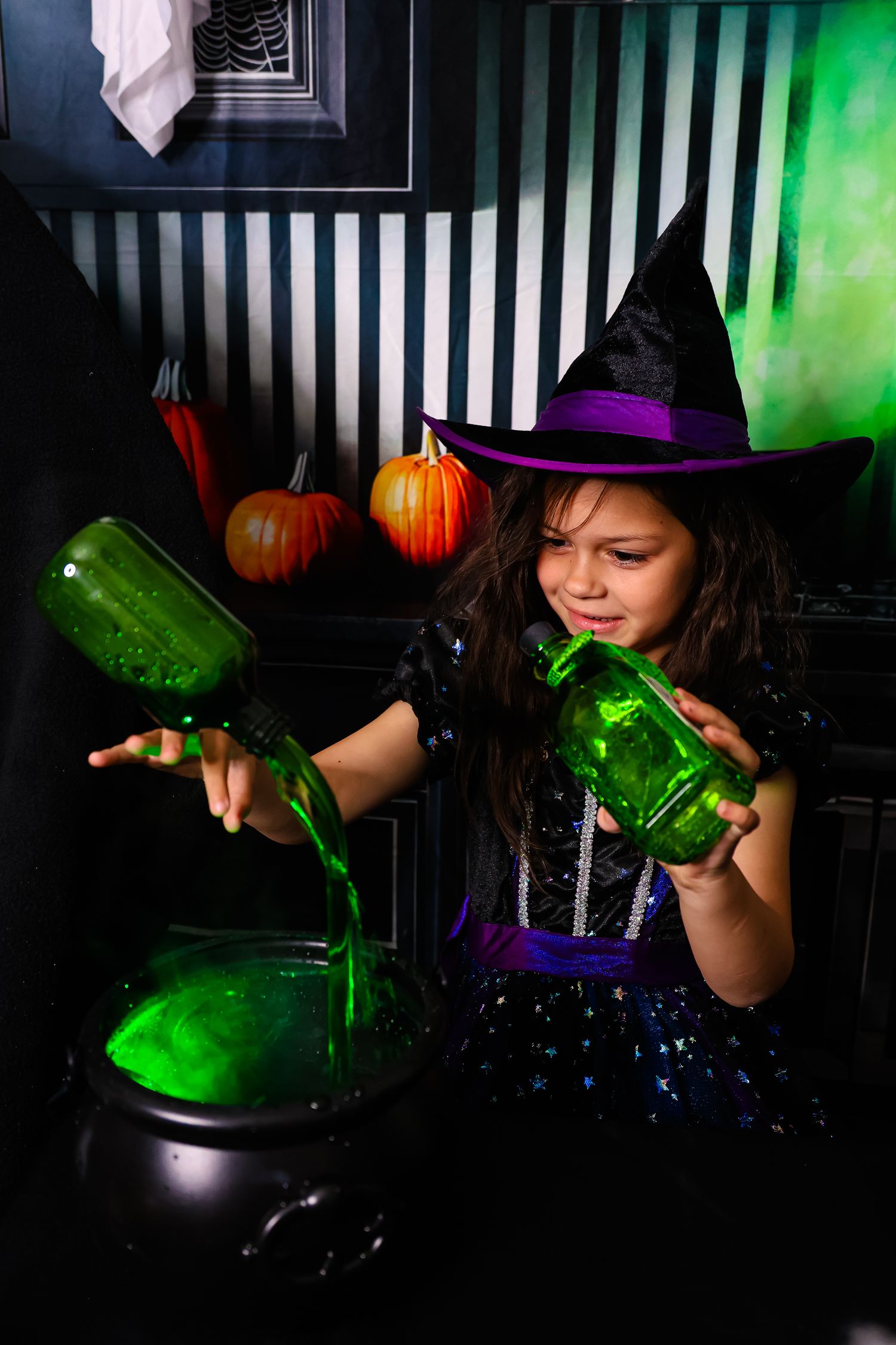 Girl in witch costume pouring green potion into cauldron with Halloween pumpkins in background.