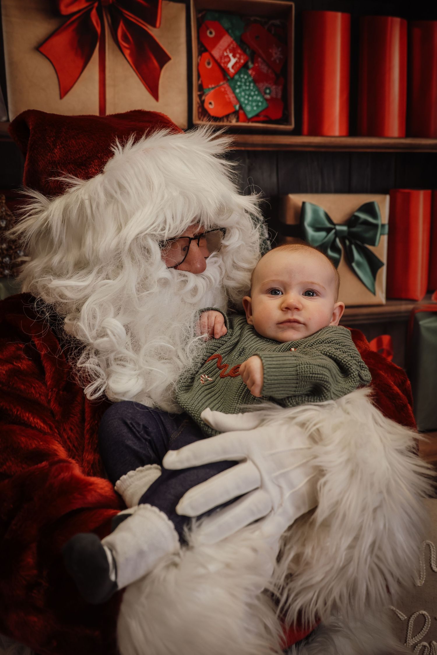 A baby in a green knit sweater sitting on Santa Claus's lap surrounded by Christmas gifts and decorations.