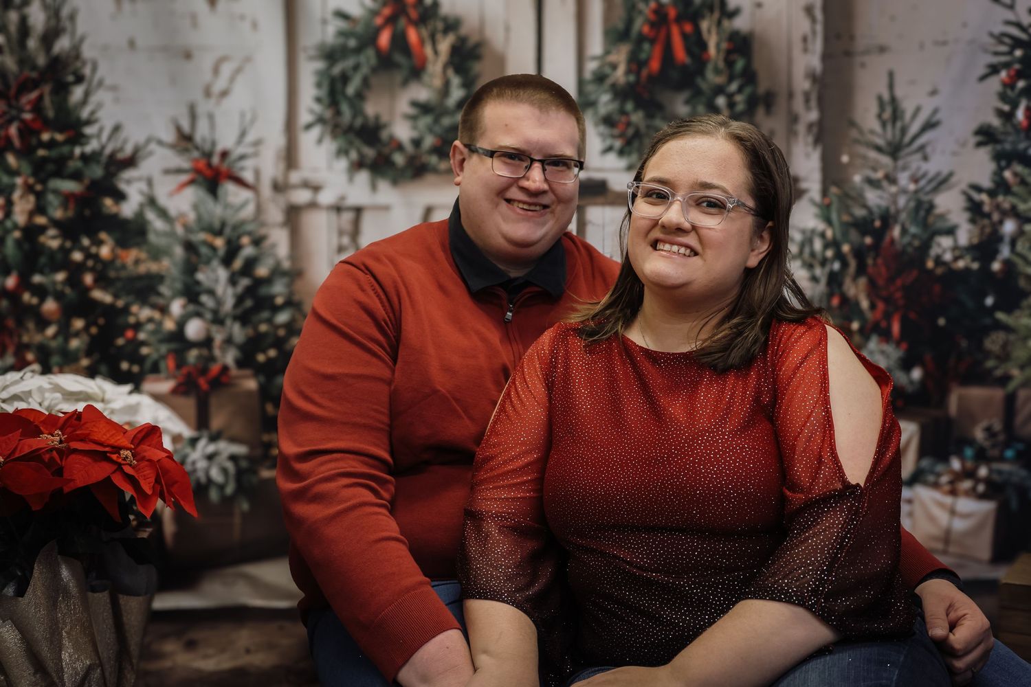 Couple in matching red sweaters smiling together in front of festive Christmas tree holiday photo backdrop.