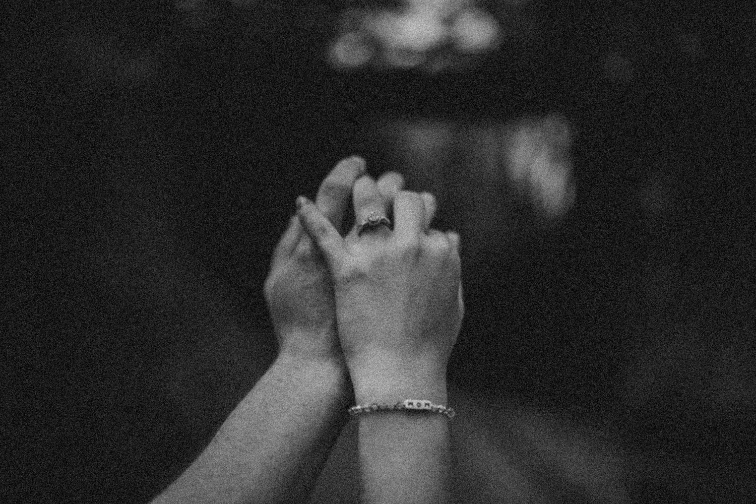 Black and white photo of two hands intertwined, one wearing a bracelet, against a dark blurred background.