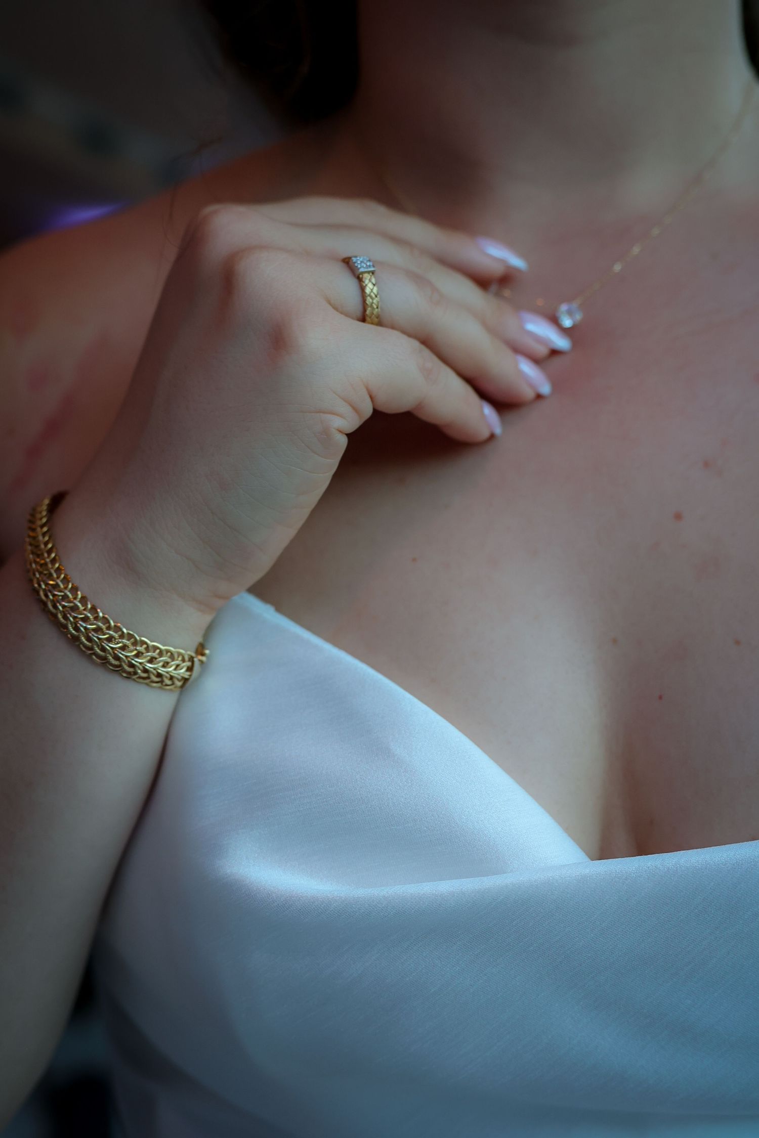 Close-up of a bride wearing a gold chain bracelet, diamond ring, and white dress, resting her hand on her chest.