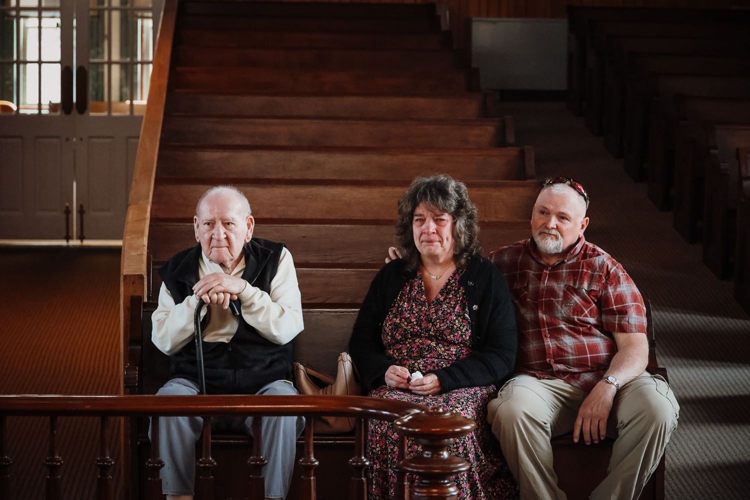 Three people sitting together on a wooden bench near a staircase, posing for a portrait indoors.