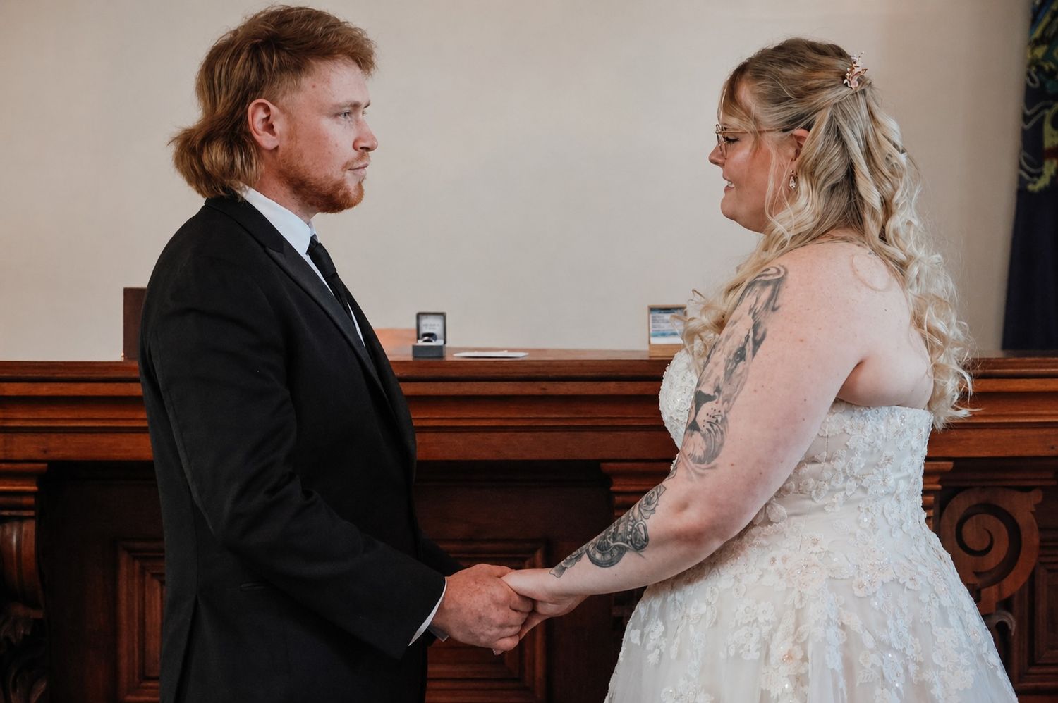 Bride and groom holding hands during wedding ceremony, groom in black suit, bride in white lace dress with tattoo.