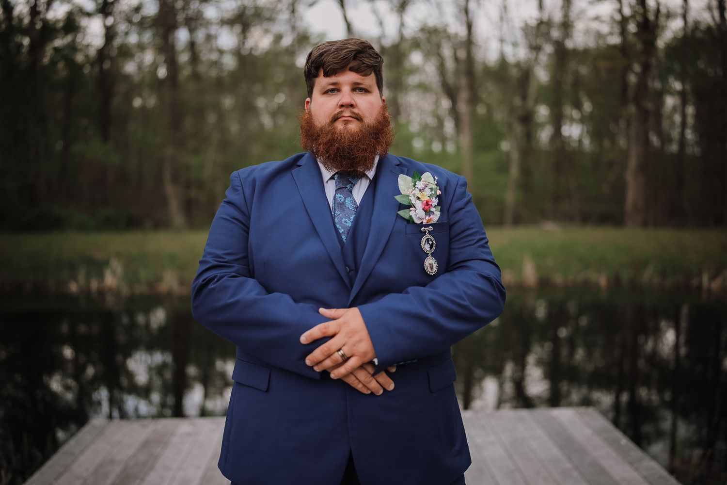 A groom in a blue suit with boutonniere stands confidently on a dock near a pond, arms crossed.