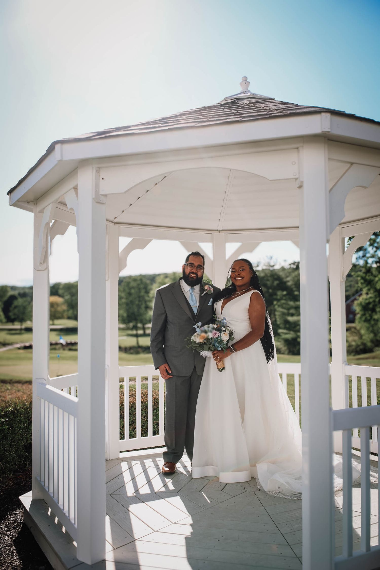 Bride and groom posing together under a white gazebo on their wedding day, with lush green golf course views behind them.
