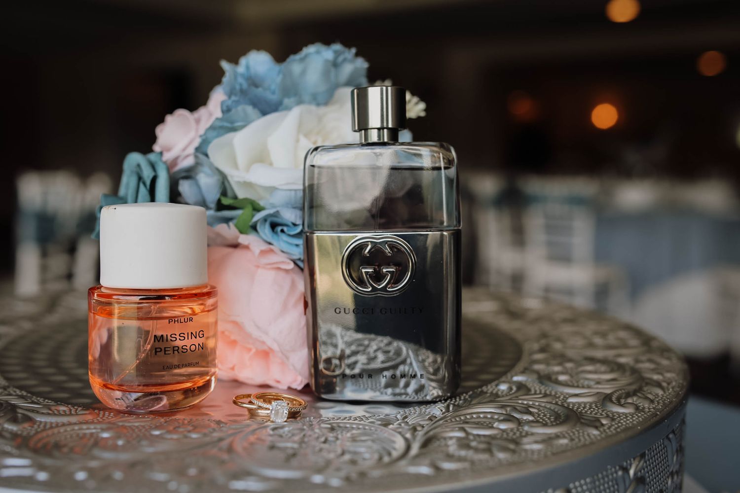 Two wedding perfume bottles, Gucci Guilty and a small orange fragrance, displayed on an ornate silver table with flowers.
