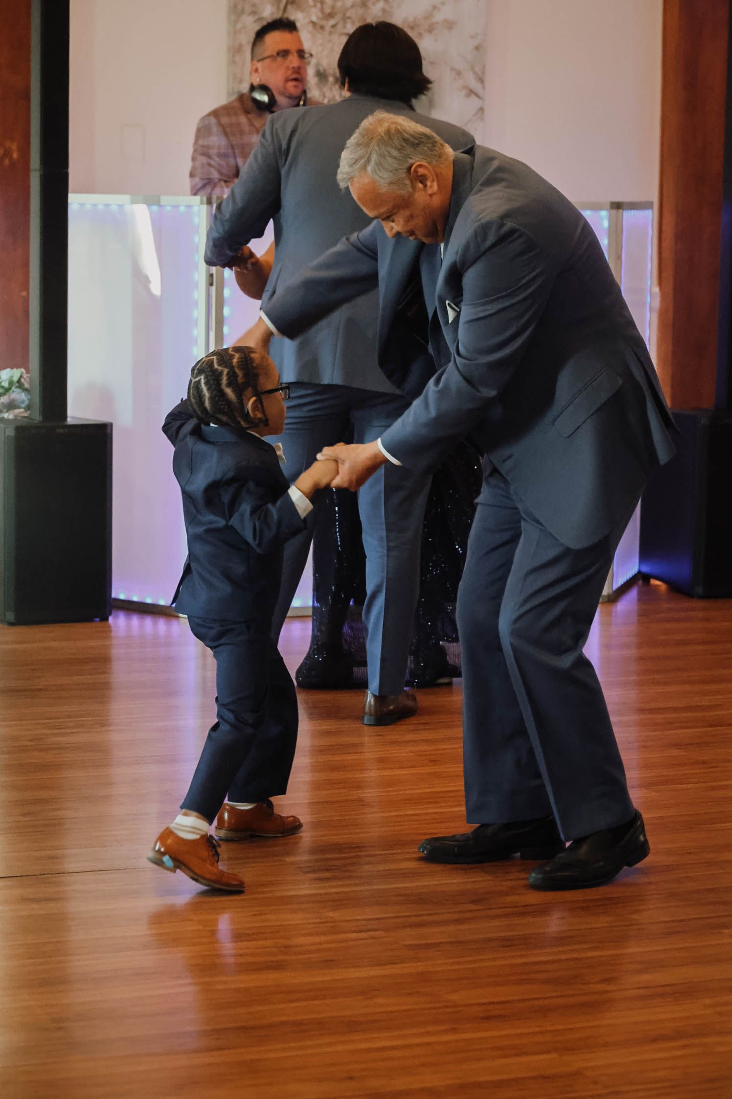 An elderly man in a blue suit dances with a young child in a matching suit on a wooden dance floor.