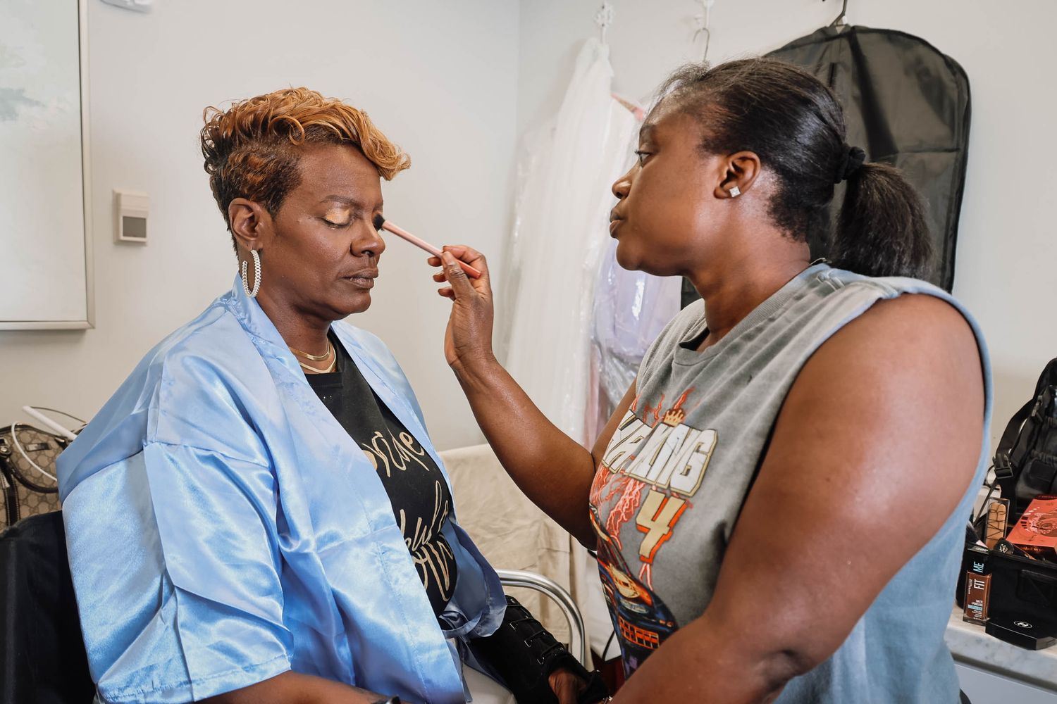 A makeup artist applies cosmetics to a woman in a blue robe in a dressing room setting.