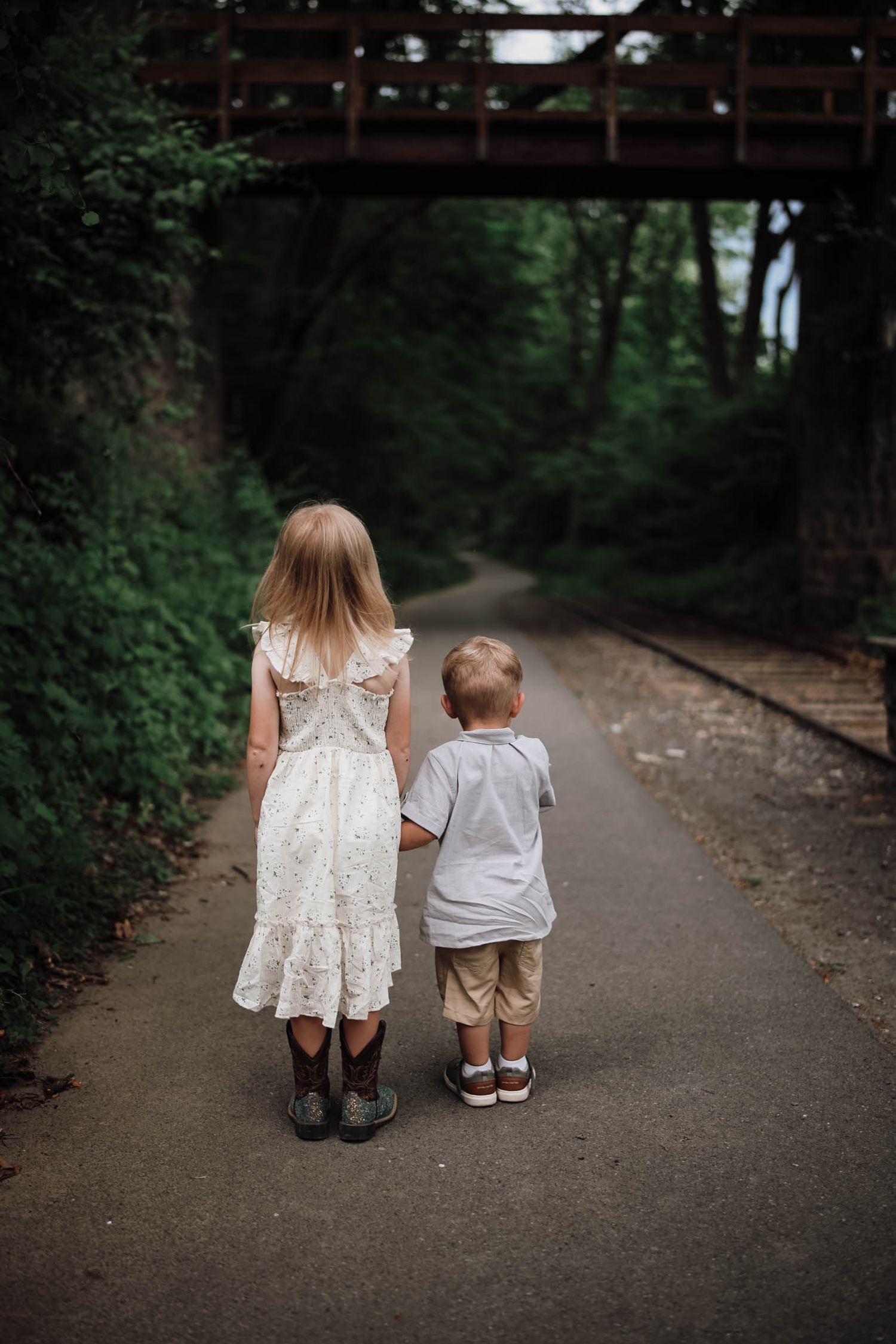 Two young children holding hands walk down a tree-lined path toward a bridge, girl in white dress and boy in gray shirt.