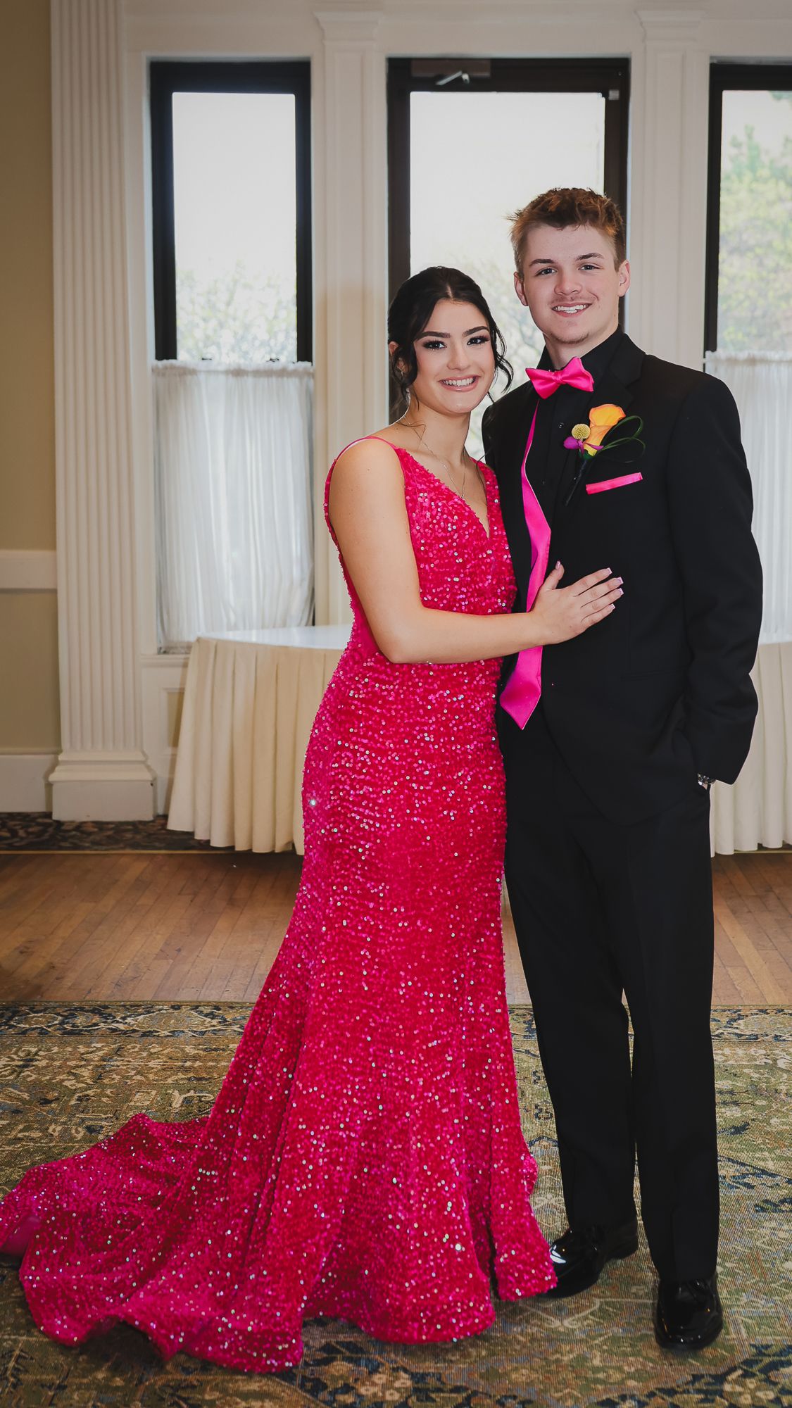 A couple poses in prom attire, she in a pink sequin gown, he in a black tux with pink bow tie.