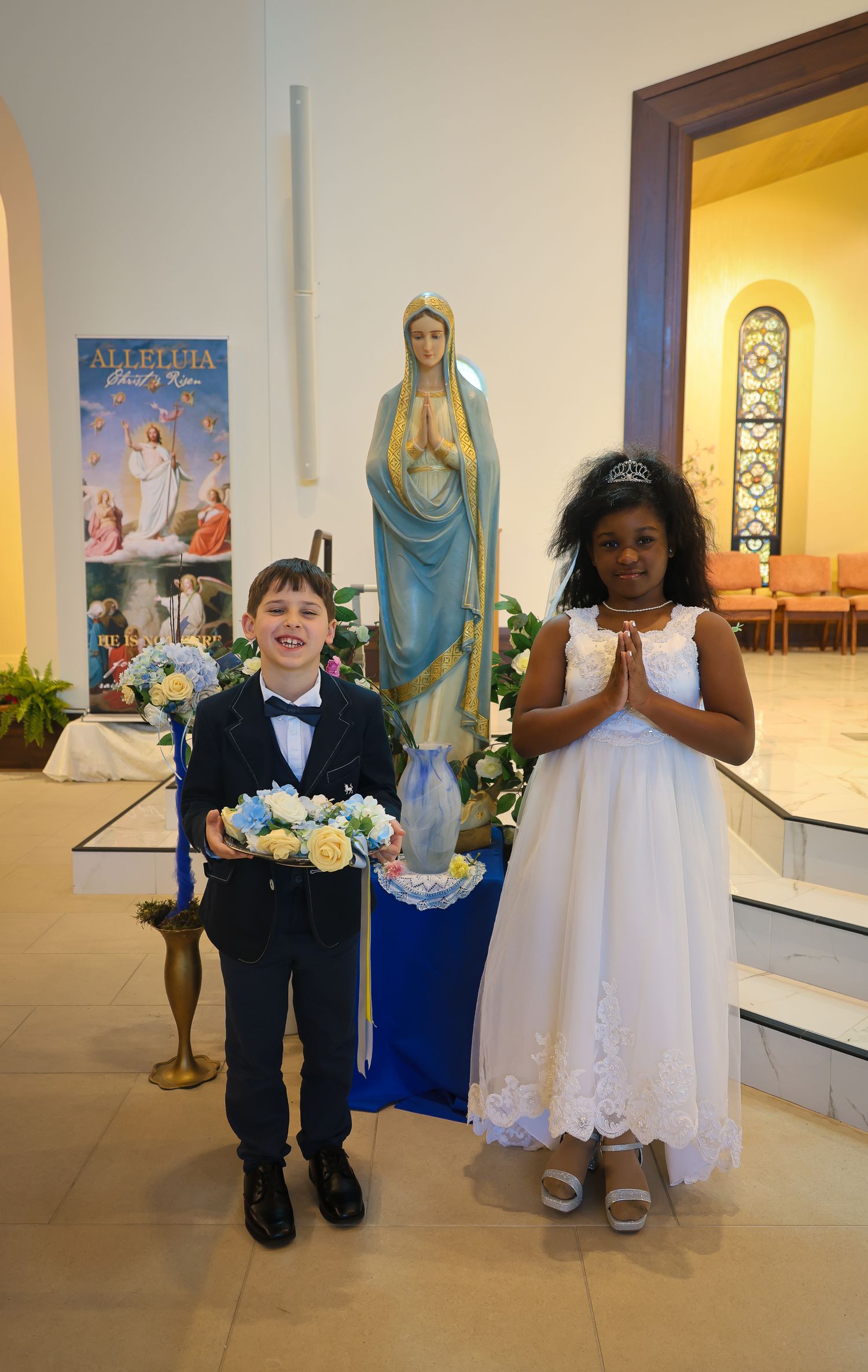 Young children in formal attire walk down a church hallway lined with religious artwork and lit displays.