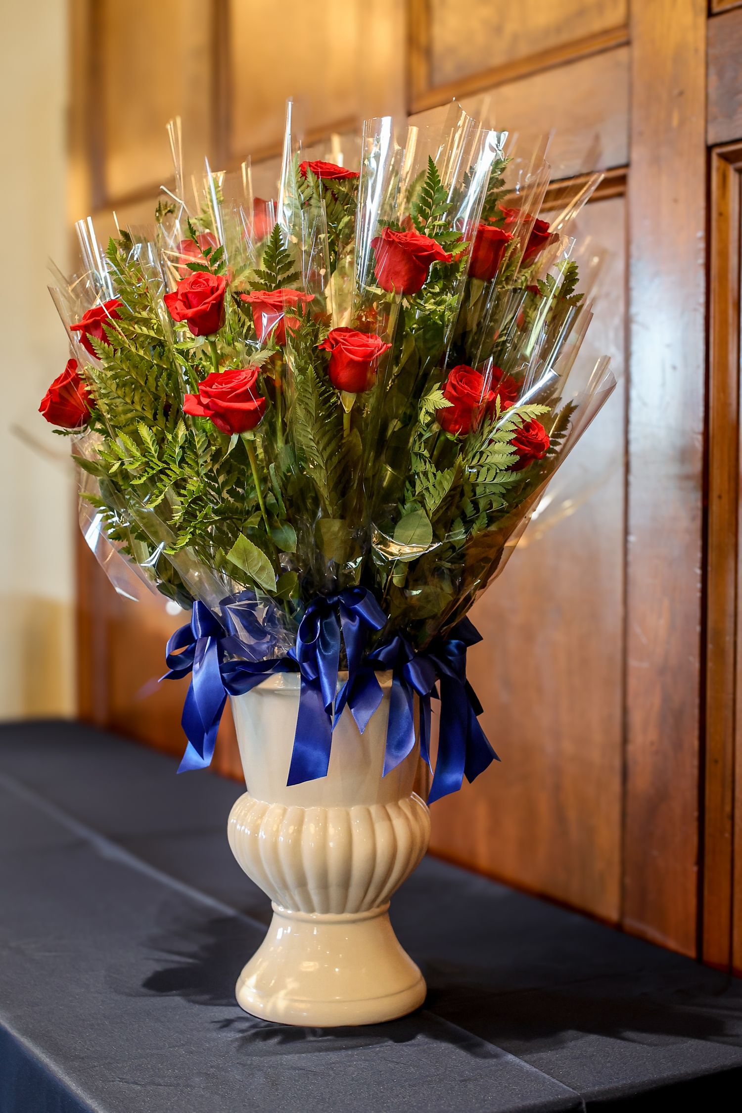 A white ceramic vase filled with red roses and greenery, decorated with a blue ribbon on a dark surface.