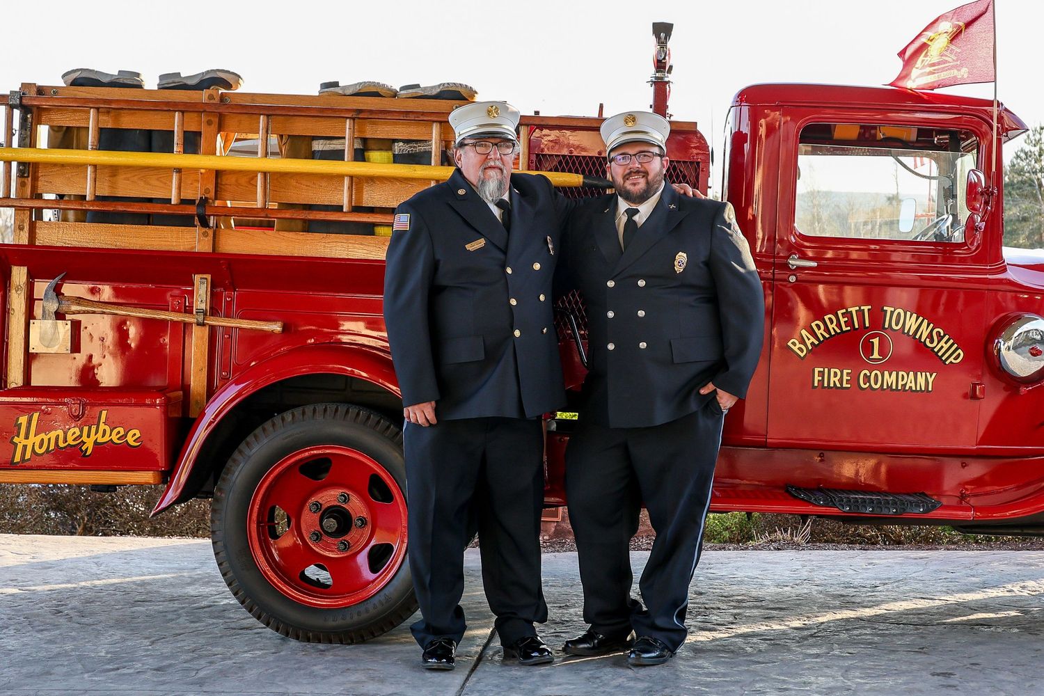 Two firefighters stand next to a vintage red fire truck with gold lettering and classic styling.
