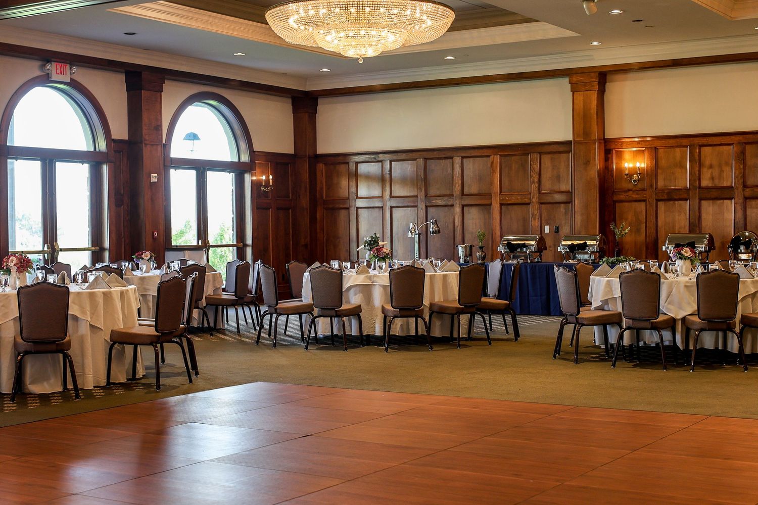 Elegant ballroom with wood paneled walls, crystal chandelier, round dining tables and large arched windows.