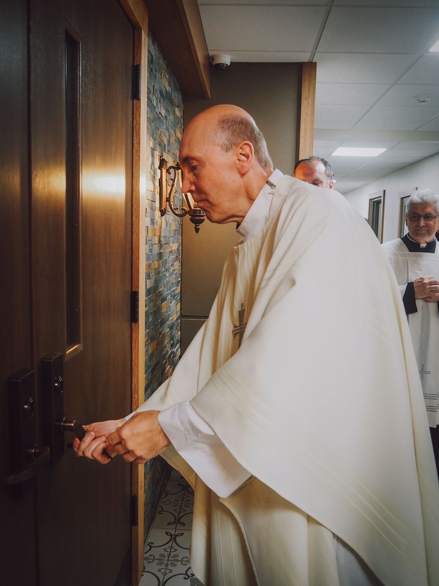 A person wearing white robes unlocks a door in a hallway with wood paneling and fluorescent lighting.