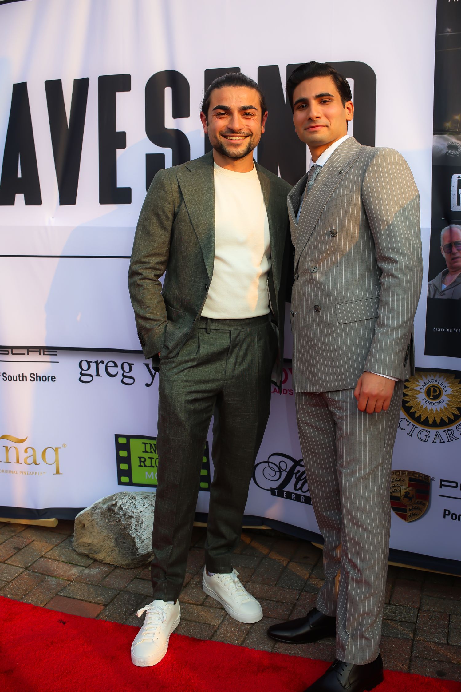 Two people in contrasting outfits pose on a red carpet at an event, with one wearing white sneakers and the other in pinstripes.