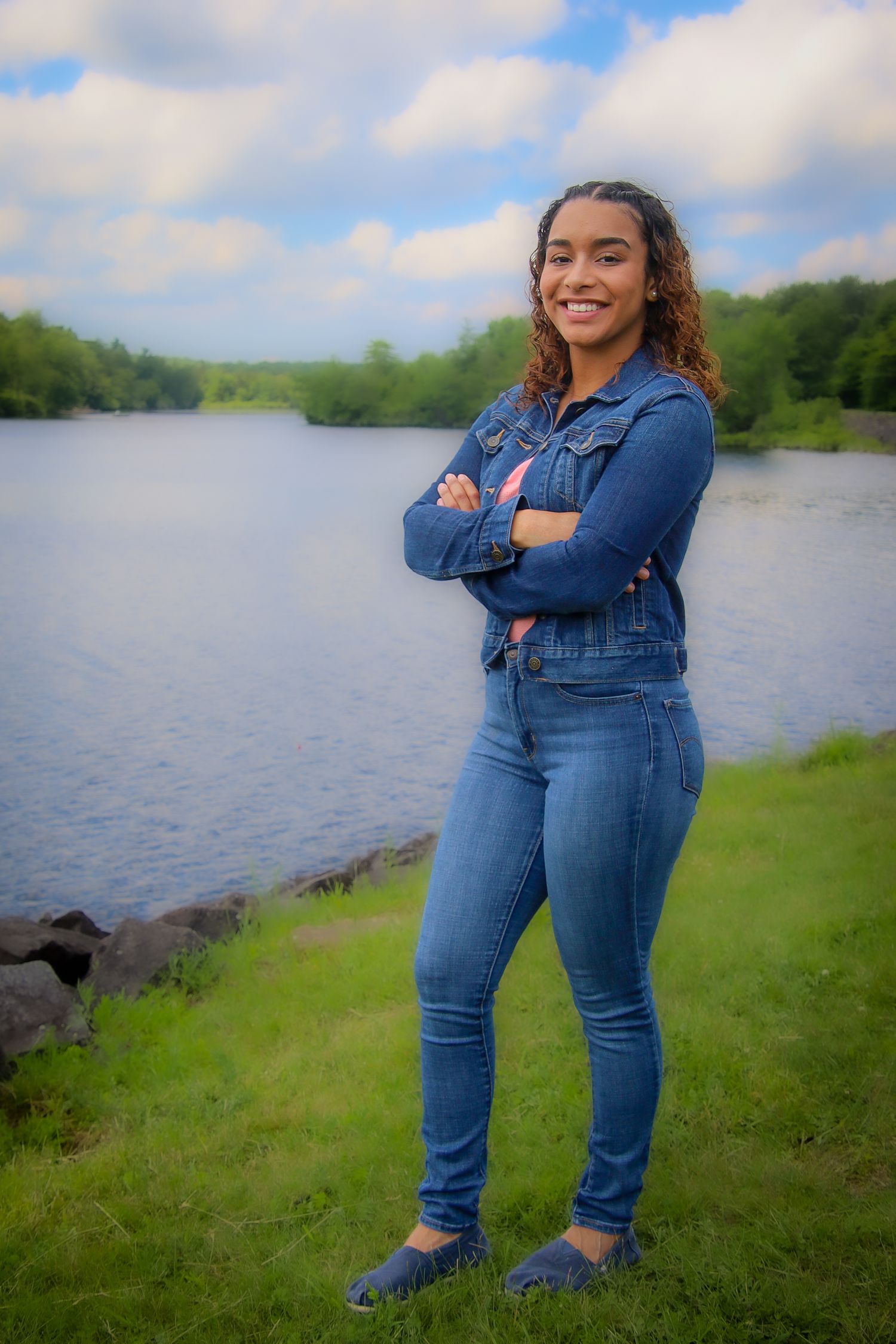 Person in double denim outfit stands confidently by a serene lake on a cloudy day with green surroundings.