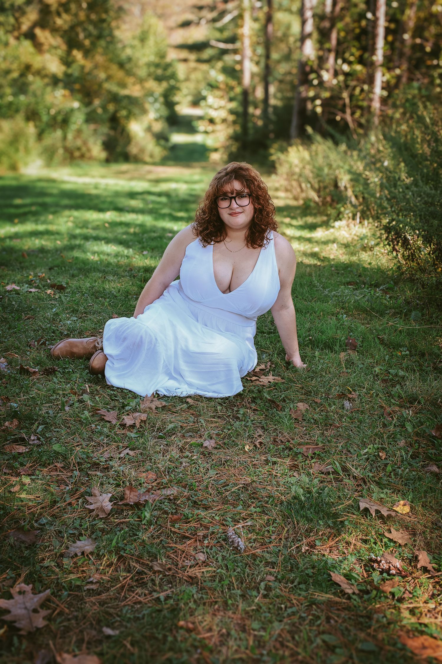 A person in a flowing white dress reclines on grass and autumn leaves in a sunlit wooded setting.