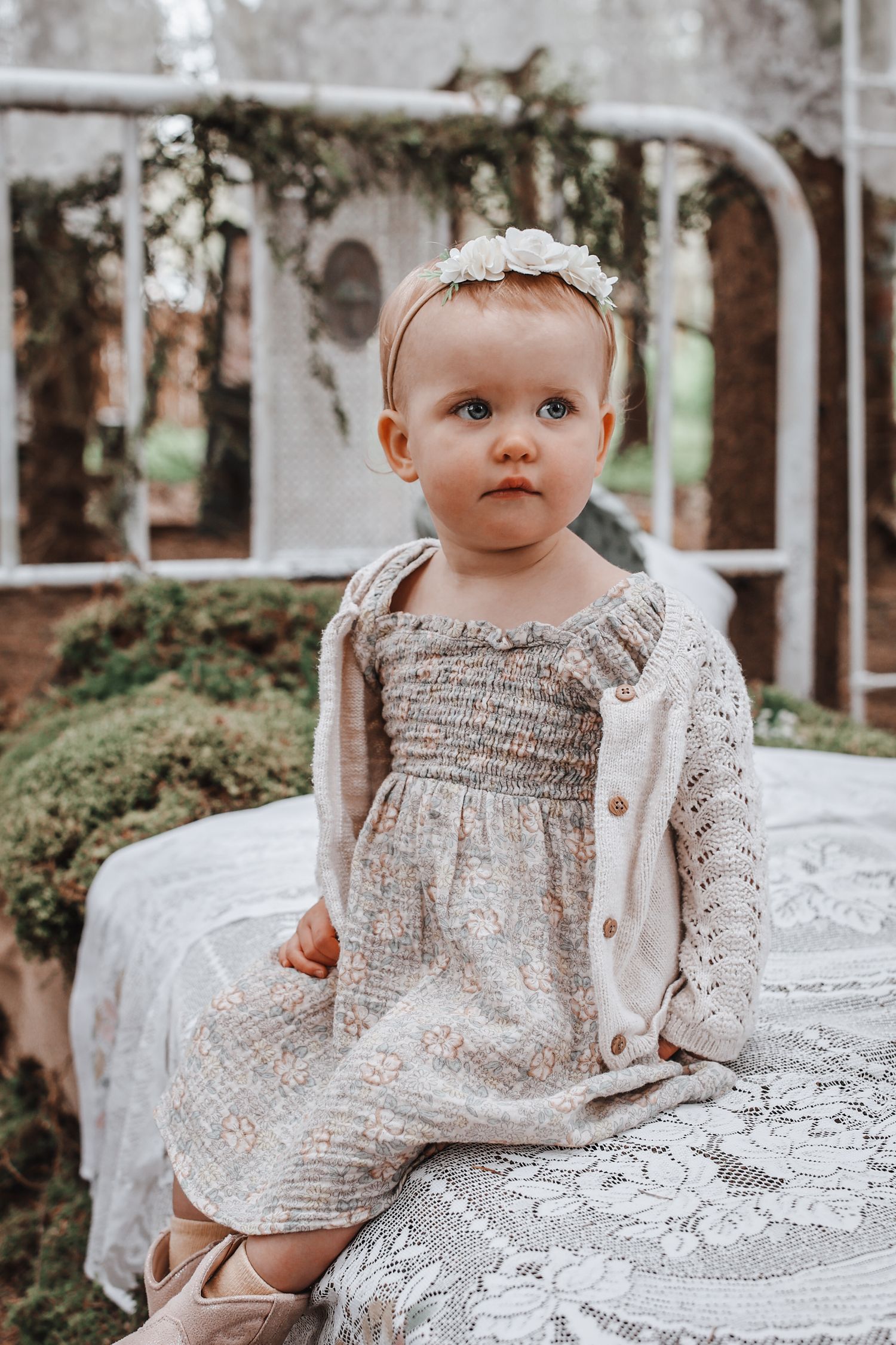 Baby wearing vintage-style lace dress sits on white lace tablecloth in outdoor garden setting.