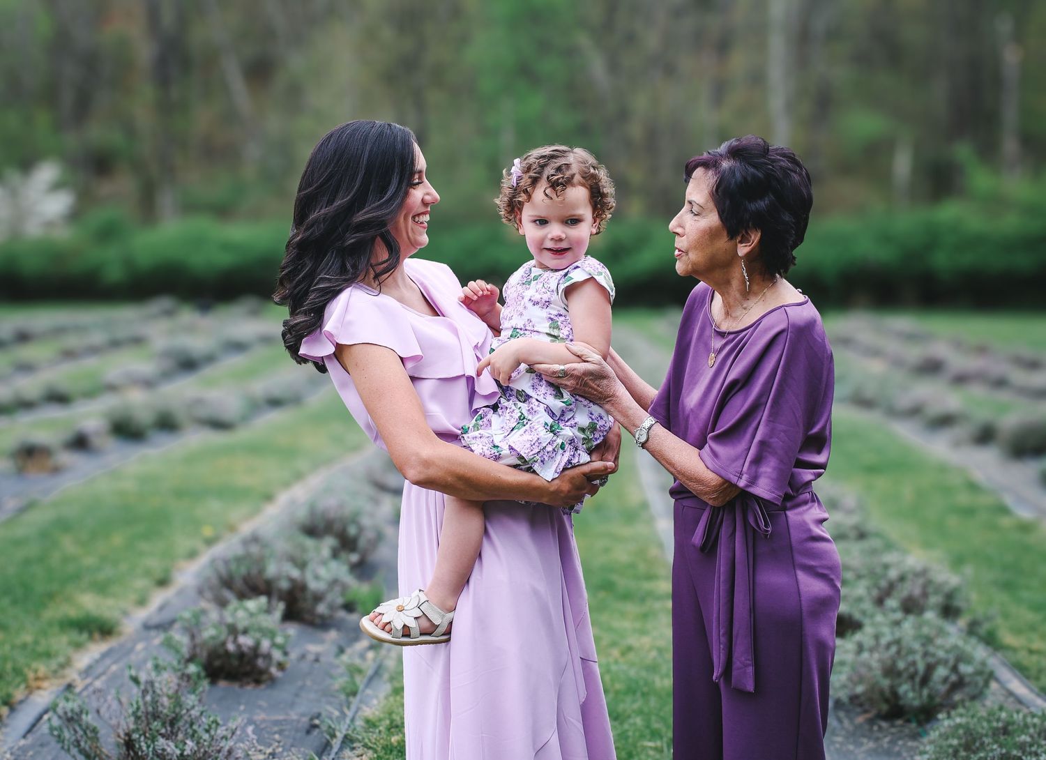 Three generations share a tender moment together in a lavender field wearing coordinating purple and pink dresses.