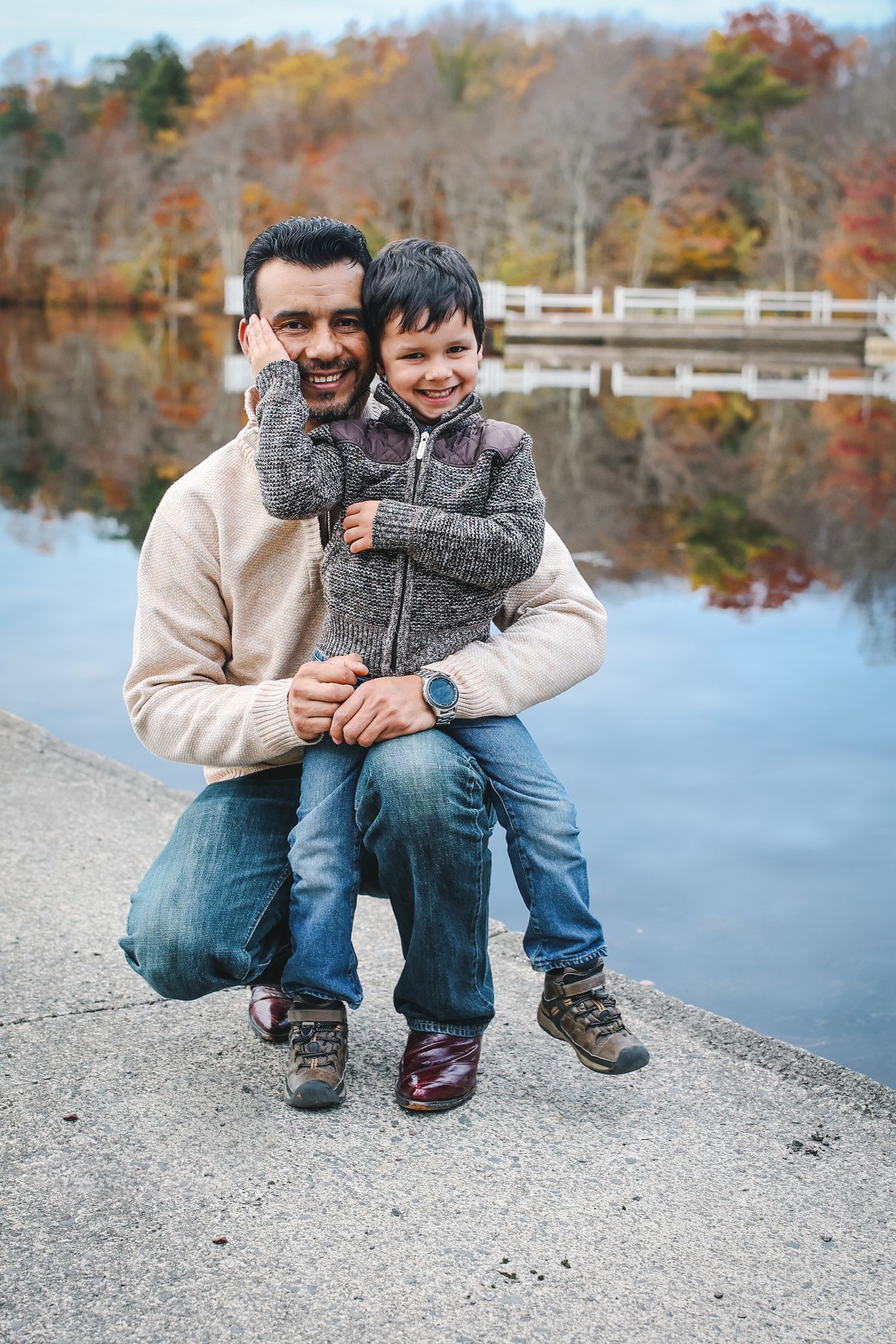 Two people sitting close together on a rocky shore by a lake with autumn trees reflected in the calm water.