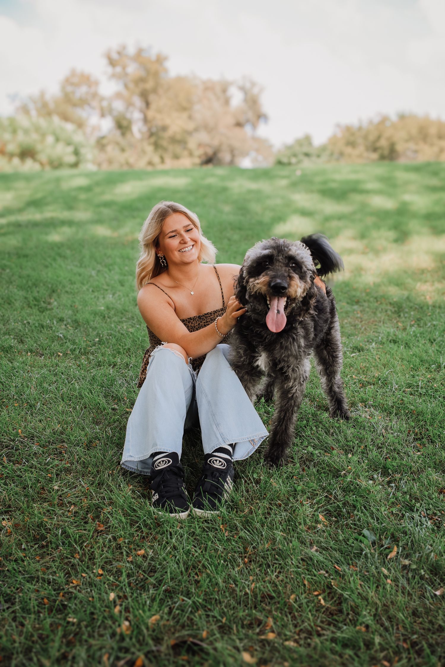 A large shaggy black dog and someone sit together on green grass in a sunny park setting.