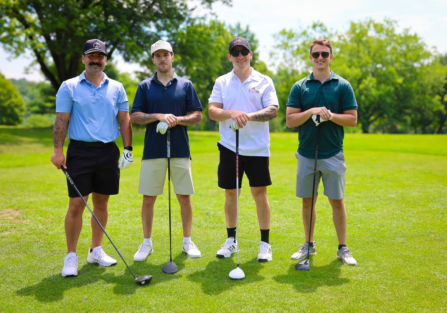 Four golfers standing together with clubs on a sunny green golf course surrounded by trees.