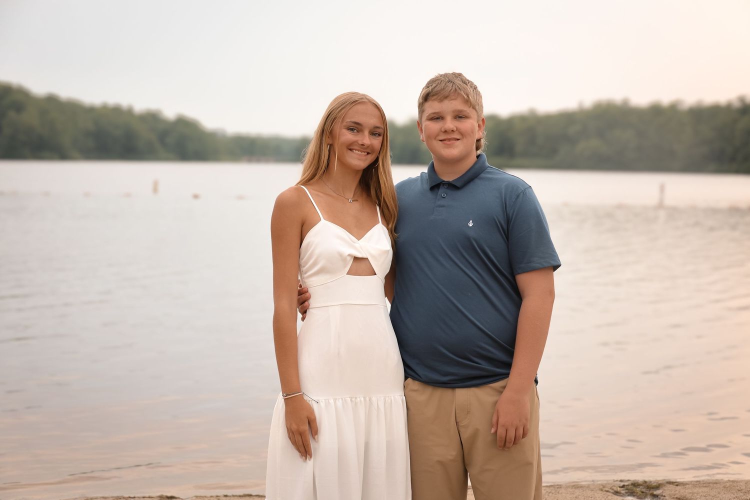A young couple stands together smiling by a serene lake at sunset, she in a white dress and he in a blue polo.