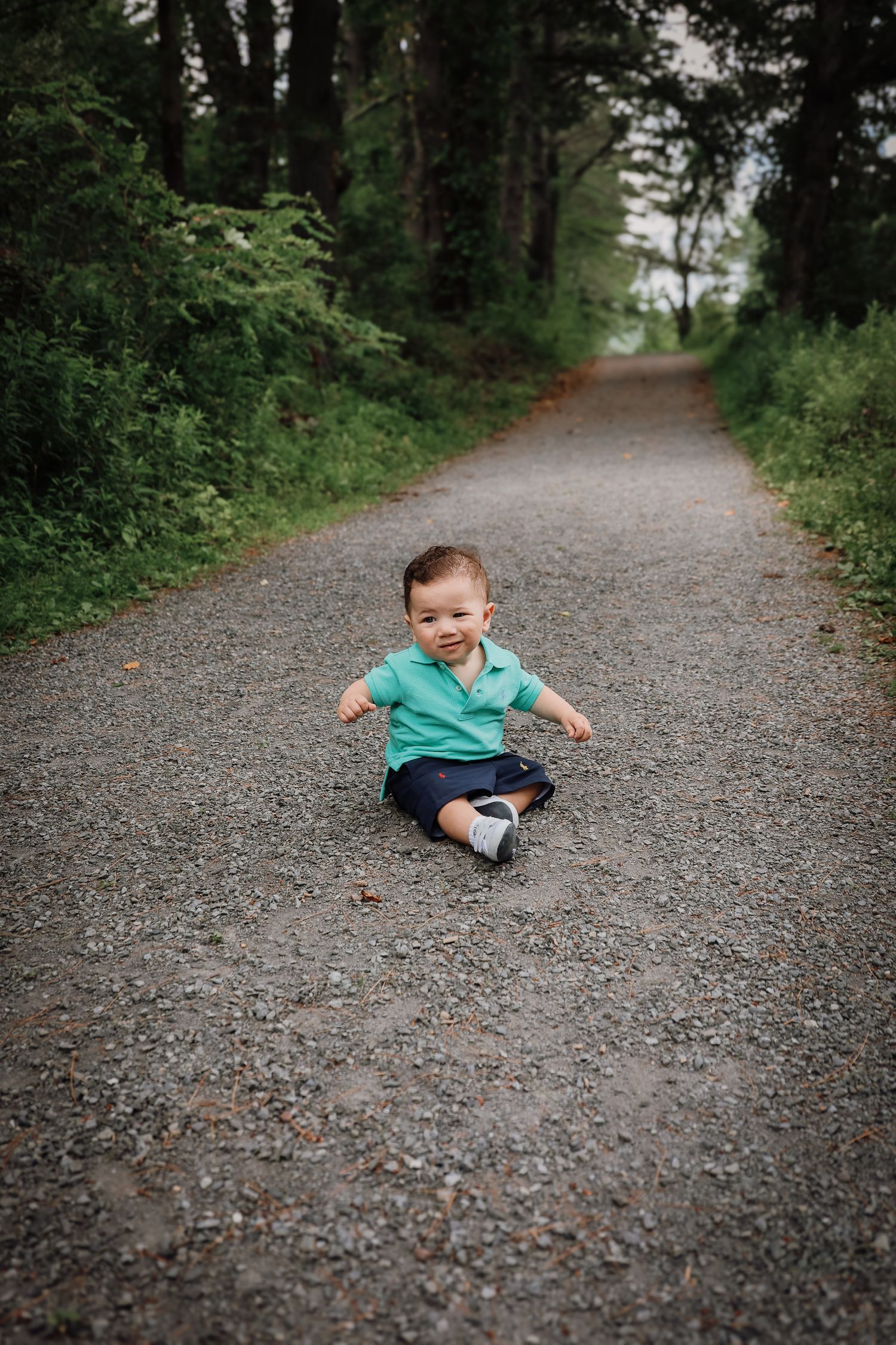 Small child in turquoise shirt sits on a gravel path surrounded by lush green forest.