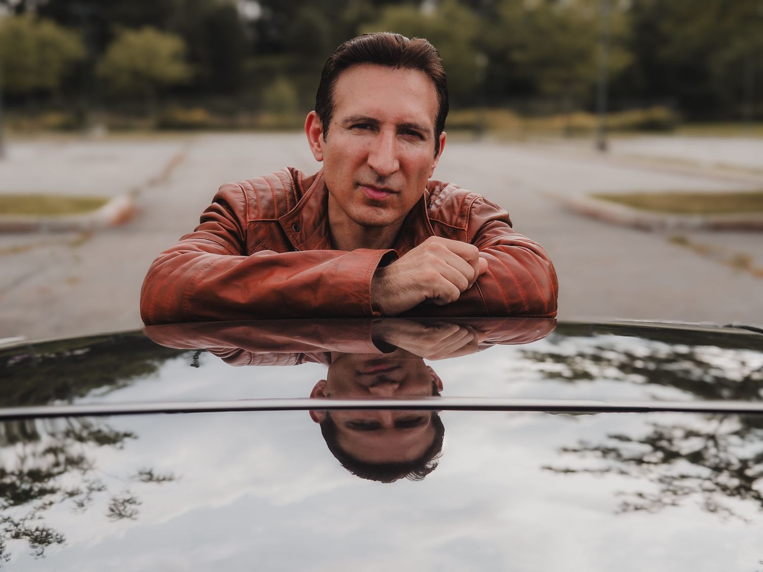 Person wearing brown leather jacket leans on car hood with reflection in overcast weather.